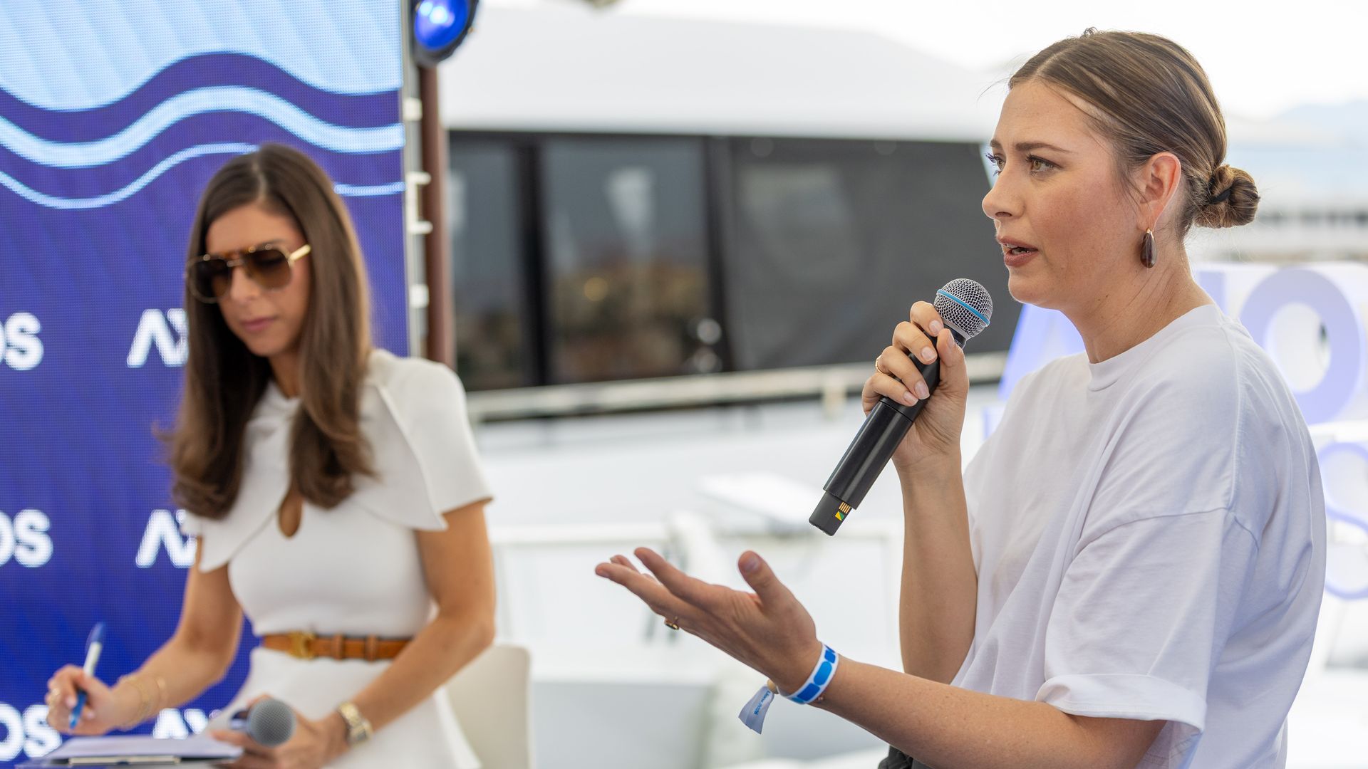 Two women at an outdoor panel: one speaks into a microphone in a white blouse with a bun; the other writes notes and wears sunglasses. A blue wave-pattern banner backdrop.