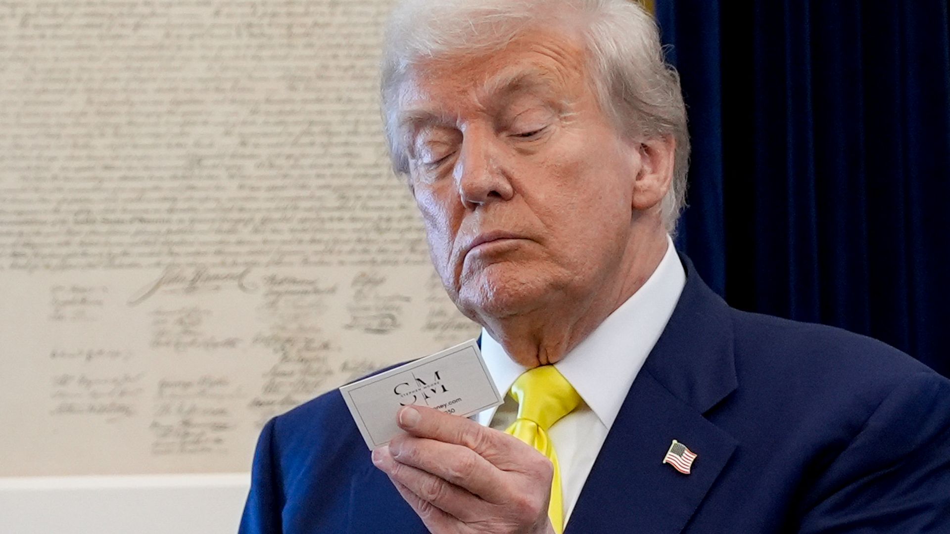 Man in navy suit with a yellow tie holding and examining a small white card closely, standing in front of a framed document with signatures.