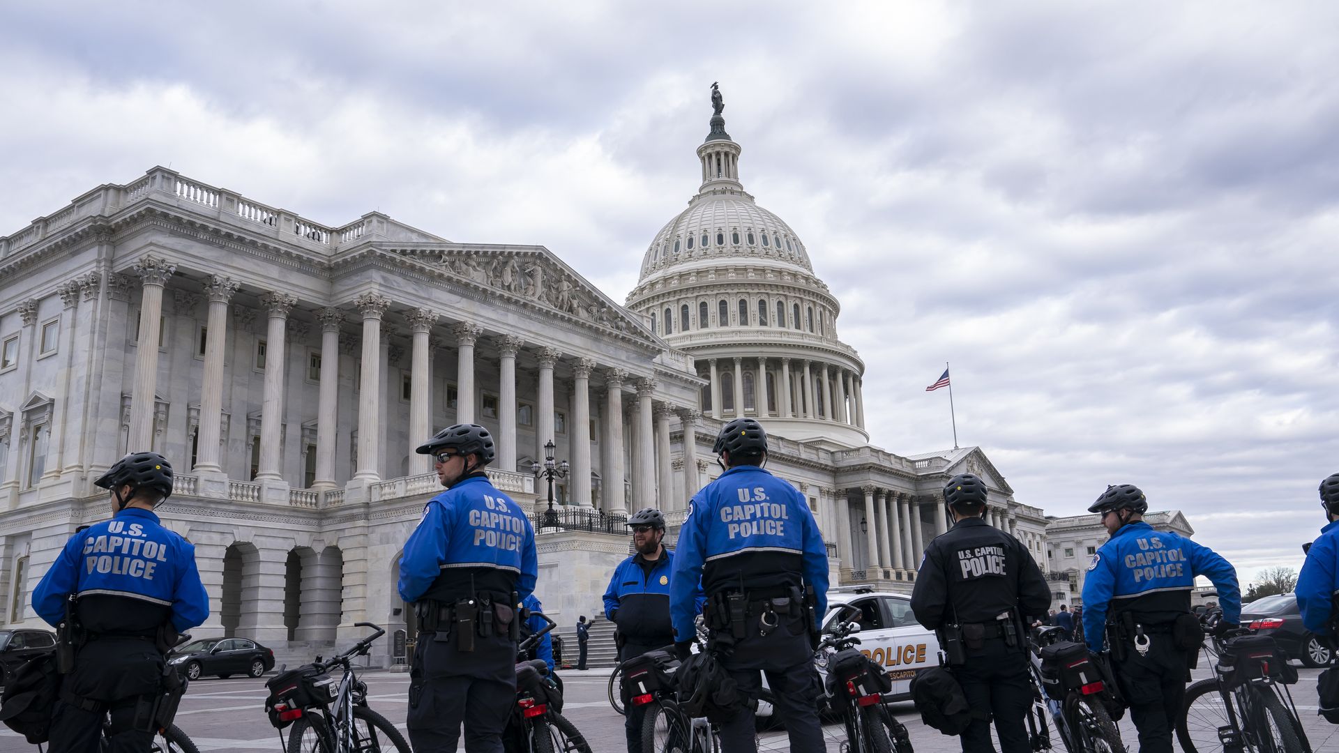A line of Capitol Police officers wearing blue "U.S. CAPITOL POLICE" jackets and holding bikes stands in front of the U.S. Capitol on a cloudy day.