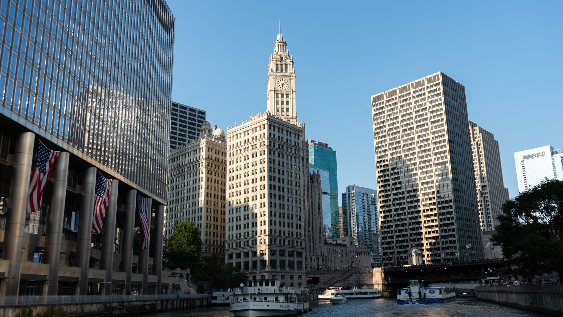 Chicago skyline along the river featuring the Wrigley Building's clock tower, glass and stone skyscrapers, American flags, boats on the water, under a clear blue sky.