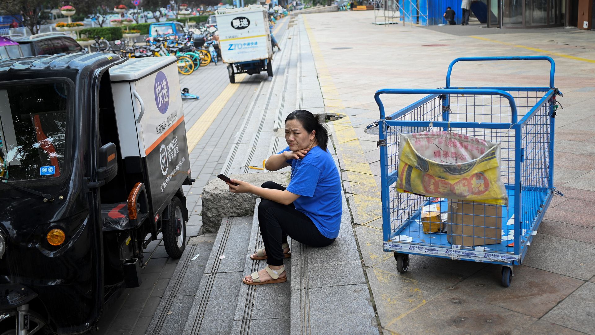 Picture of a Chinese woman sitting on steps next to a car and an empty delivery cart