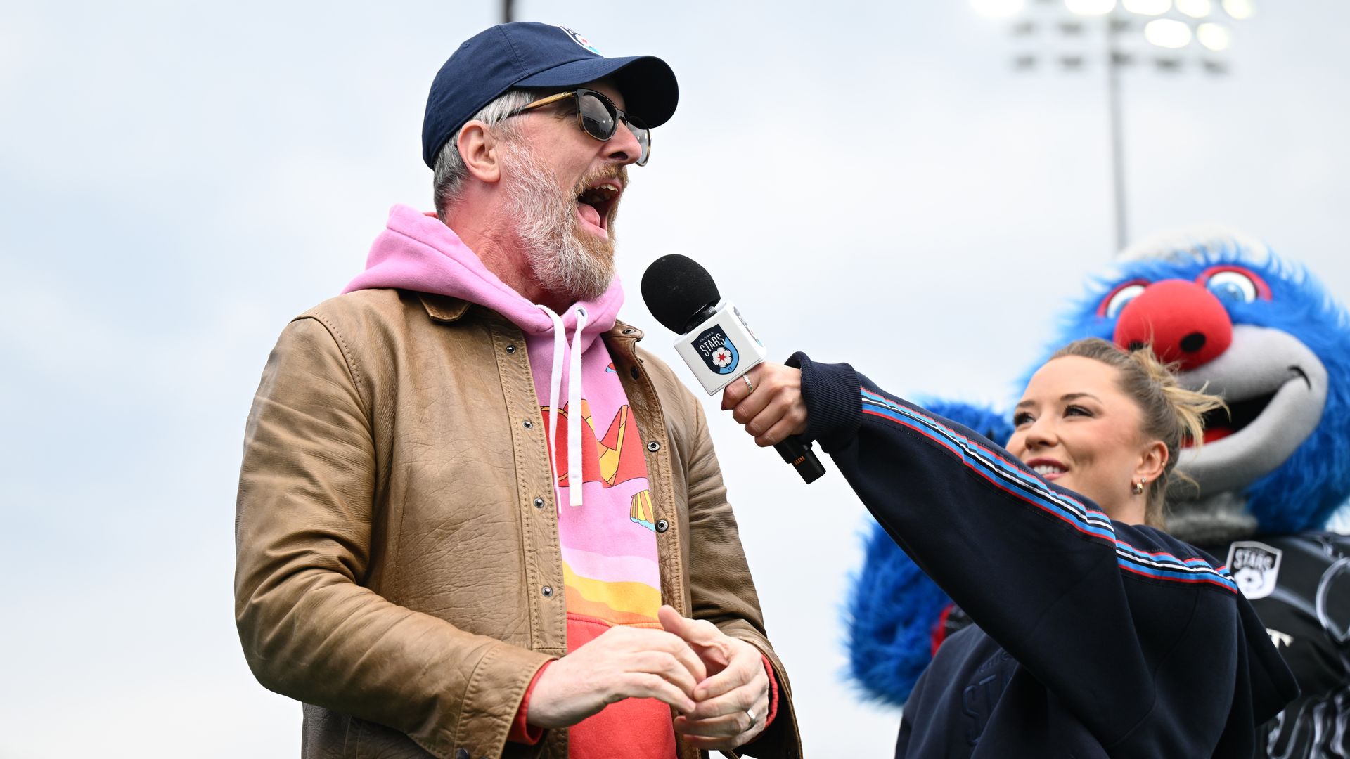 Bearded man in a pink hoodie and tan jacket speaks into a microphone, wearing sunglasses and a navy cap; a person holds the mic while a blue mascot and stadium lights loom in the background.