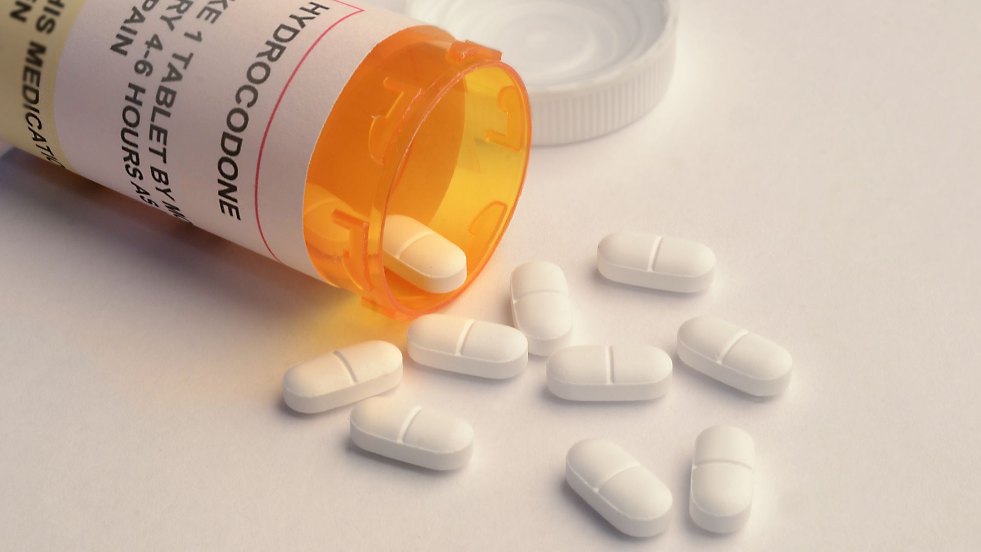 In this image, an open orange pill bottle spills small white pills onto a plain white countertop.