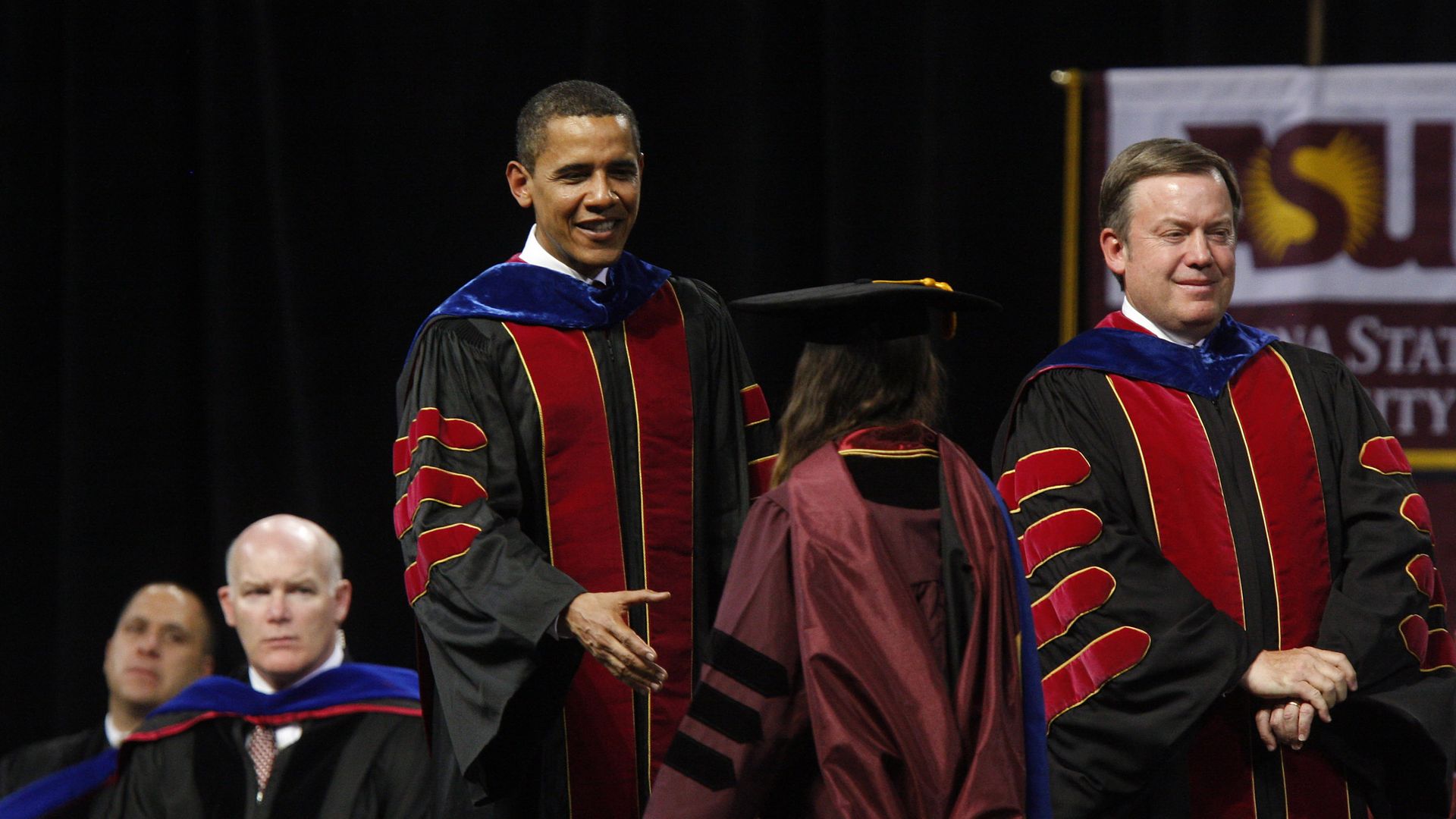 President Obama wears red and black academic robes while reaching out to shake hands with red-robed woman in a mortarboard. Another man in red and black robes stands to his left with his hands clasped in front of him. A banner for Arizona State University is in the background.