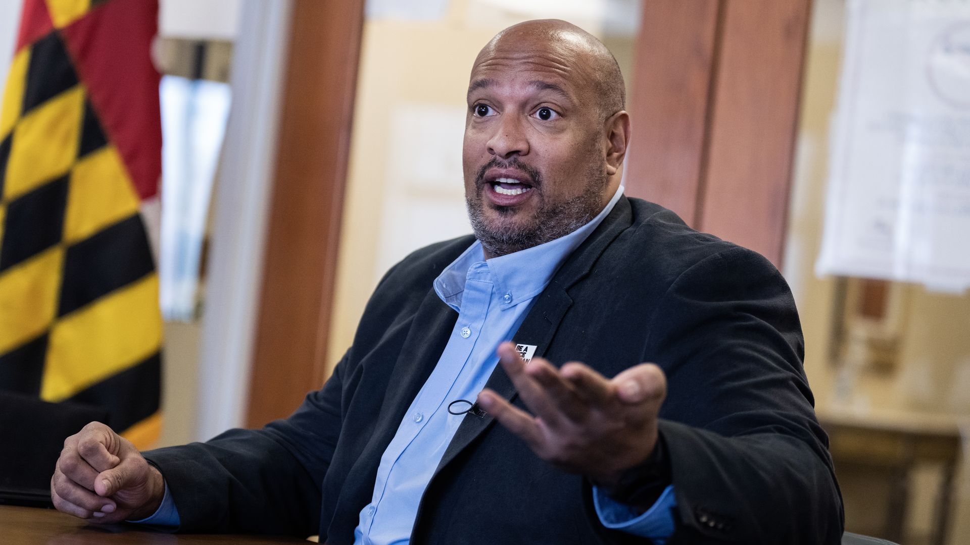 Harry Dunn, wearing a dark blue suit and blue shirt, sitting in front of a Maryland flag.
