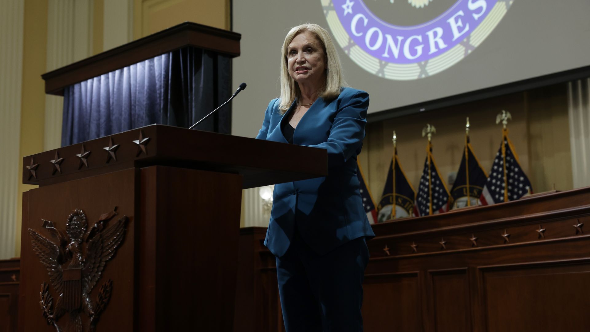 Rep. Carolyn Maloney, wearing a blue suit, speaks at a lecturn during a portrait unveiling at a House committee chamber.
