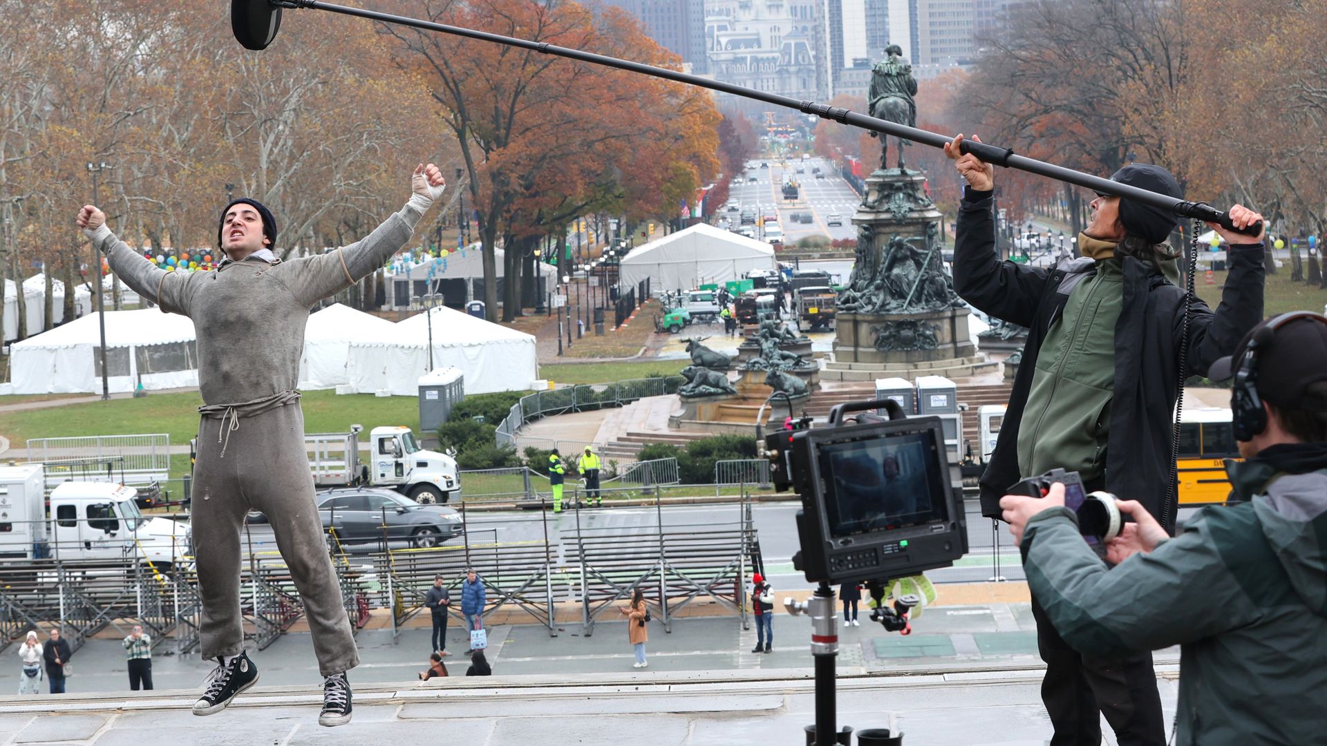 Actor wearing gray jumpsuit and black sneakers jumps with fists raised on city steps during filming, with crew holding boom mic and camera, autumn trees and cityscape in background.