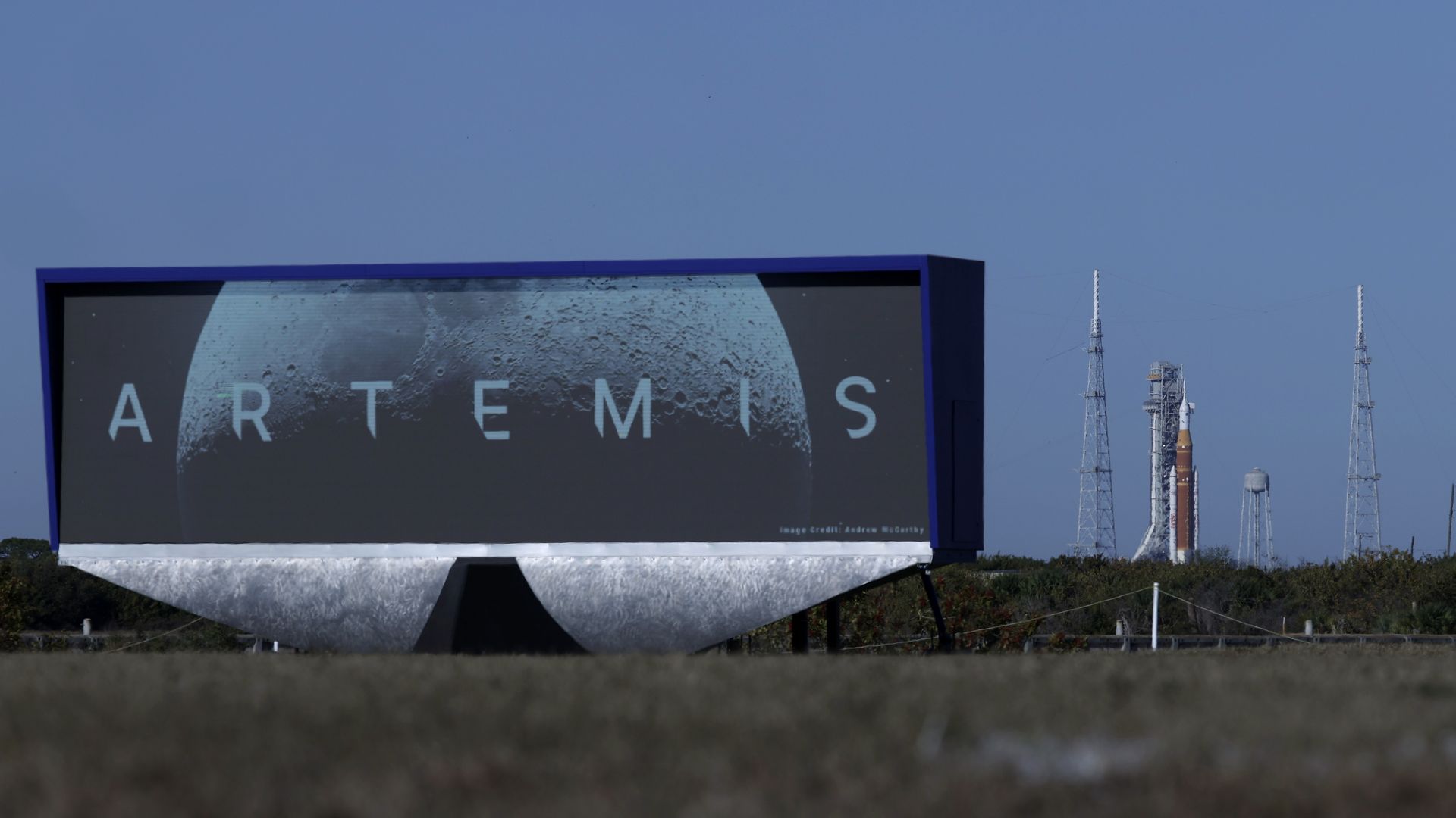 The Artemis II rocket stands on the launch stand, with a large billboard in the foreground reading: "Artemis"
