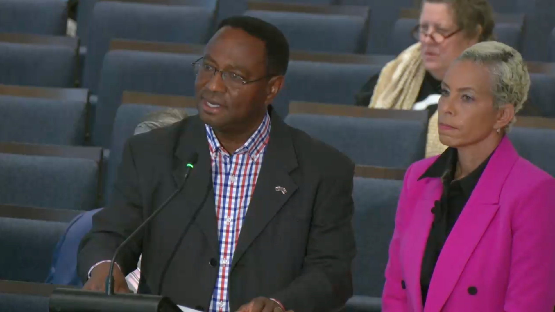 Man in black blazer and checked shirt speaking at microphone, woman in bright pink blazer and black shirt standing beside him, seated woman with glasses and scarf in background