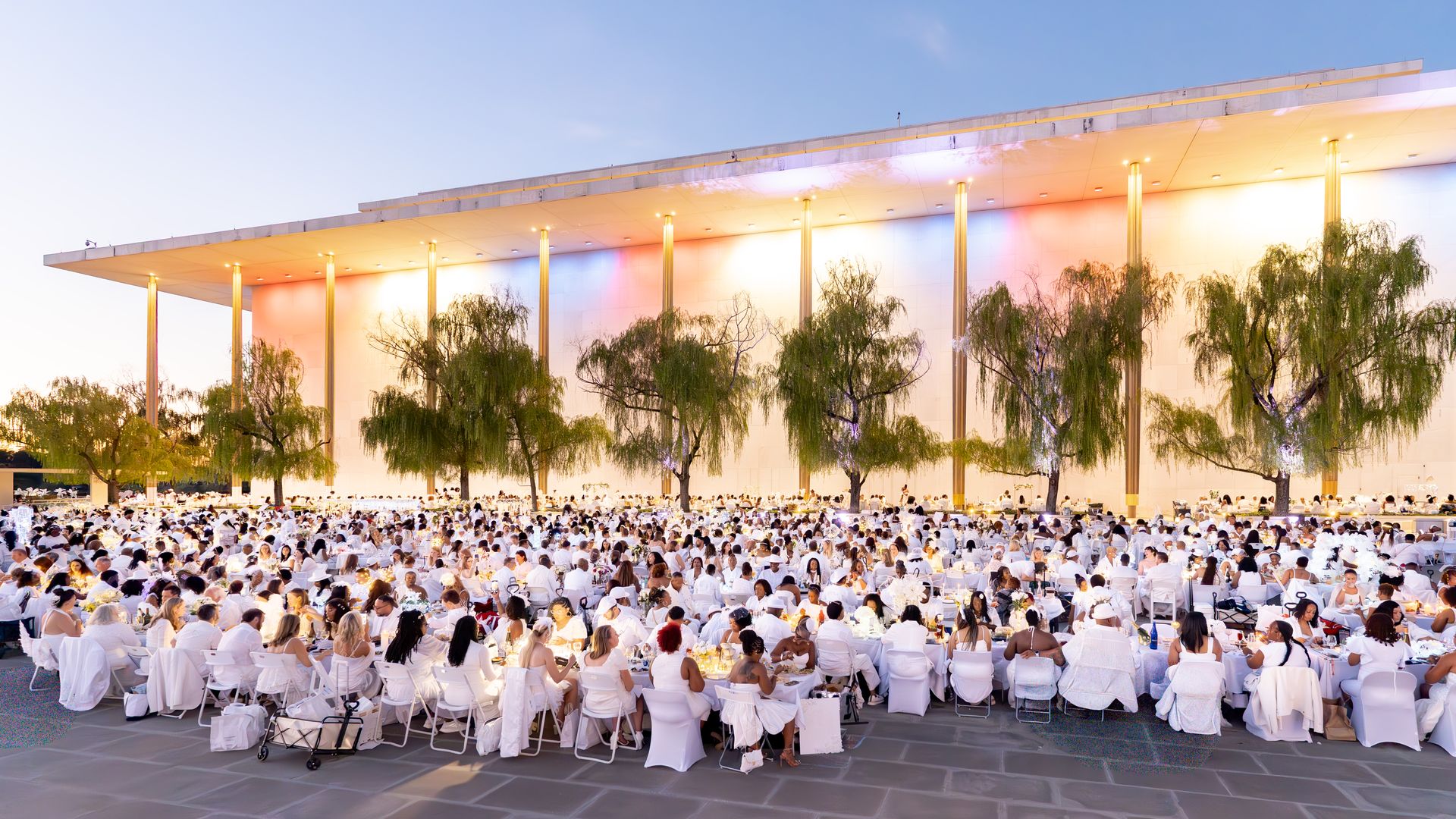 Wide outdoor event with hundreds in white, seated on white chairs under a modern columned pavilion. Warm lights glow, trees line the foreground, and a blue sky above.
