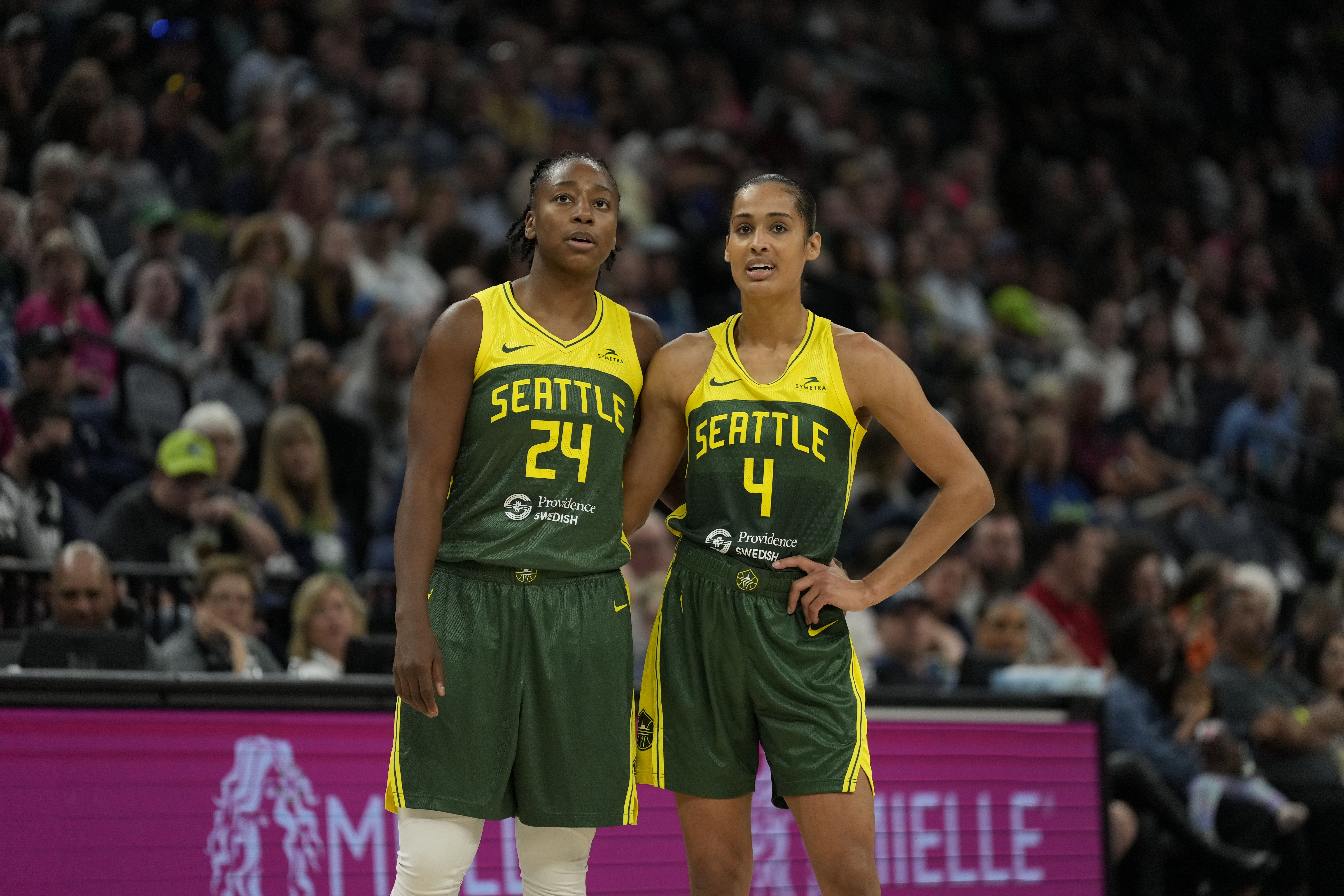 Two female basketball players in yellow and green uniforms stand closely together on the court. 