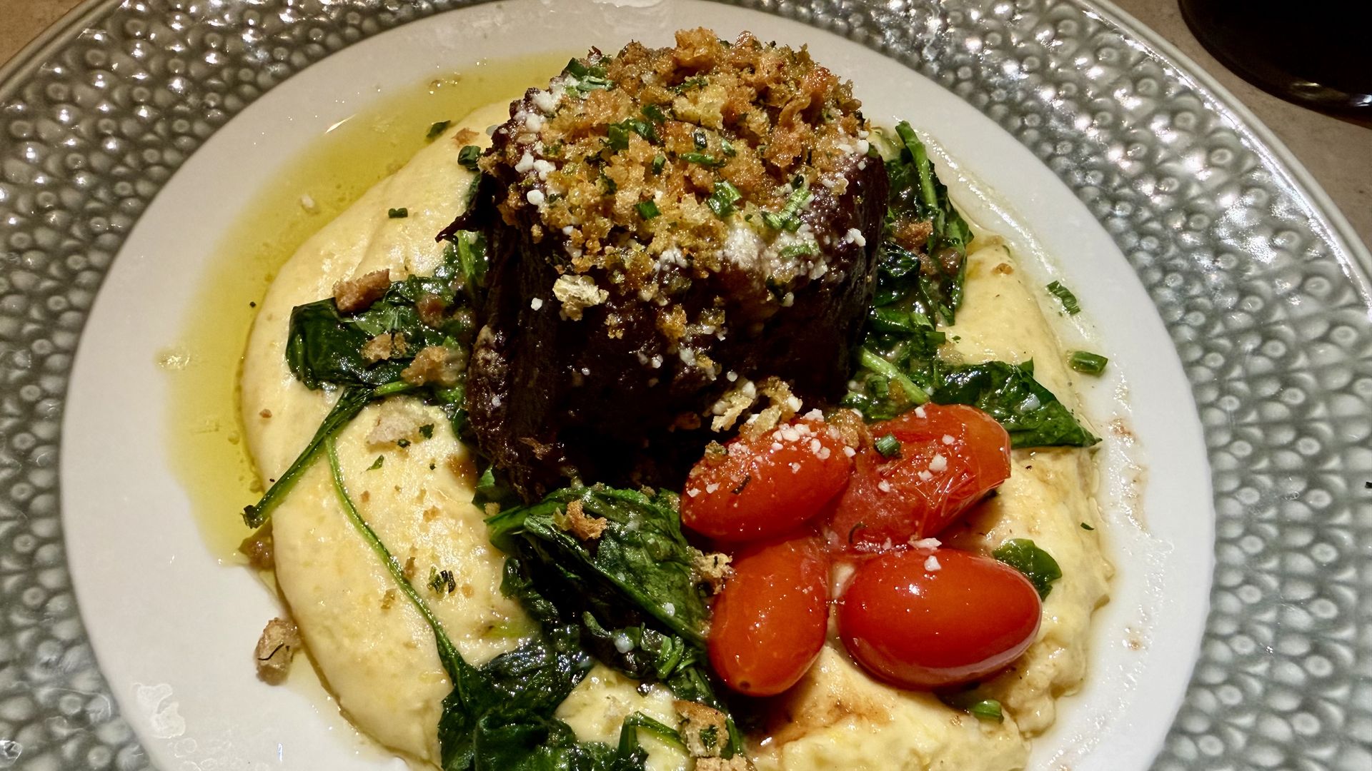 Plate with creamy polenta topped with cooked spinach, grilled cherry tomatoes, and a piece of meat covered with herb and breadcrumb crust.