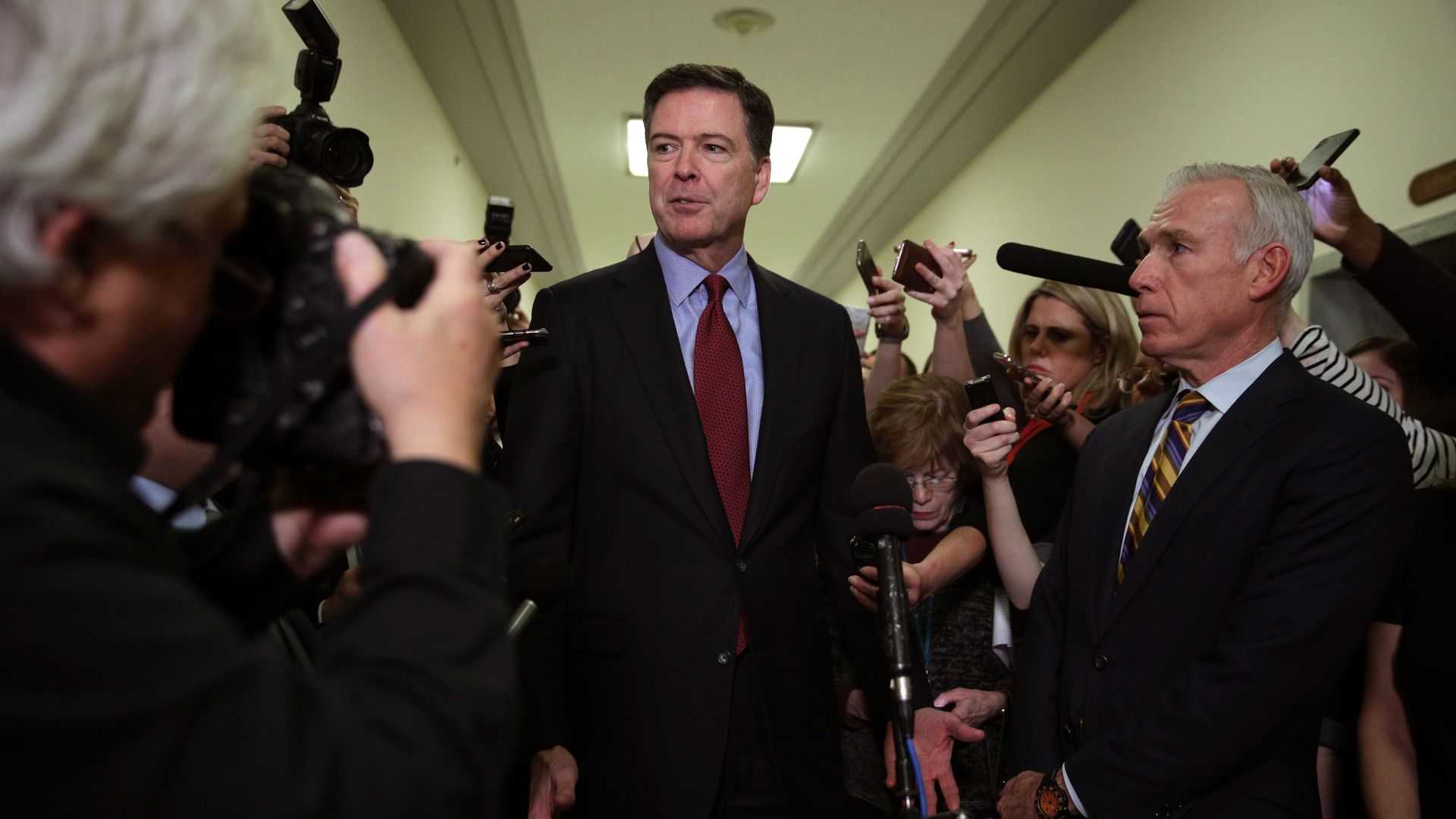 James Comey stands surrounded by reporters in a hallway.