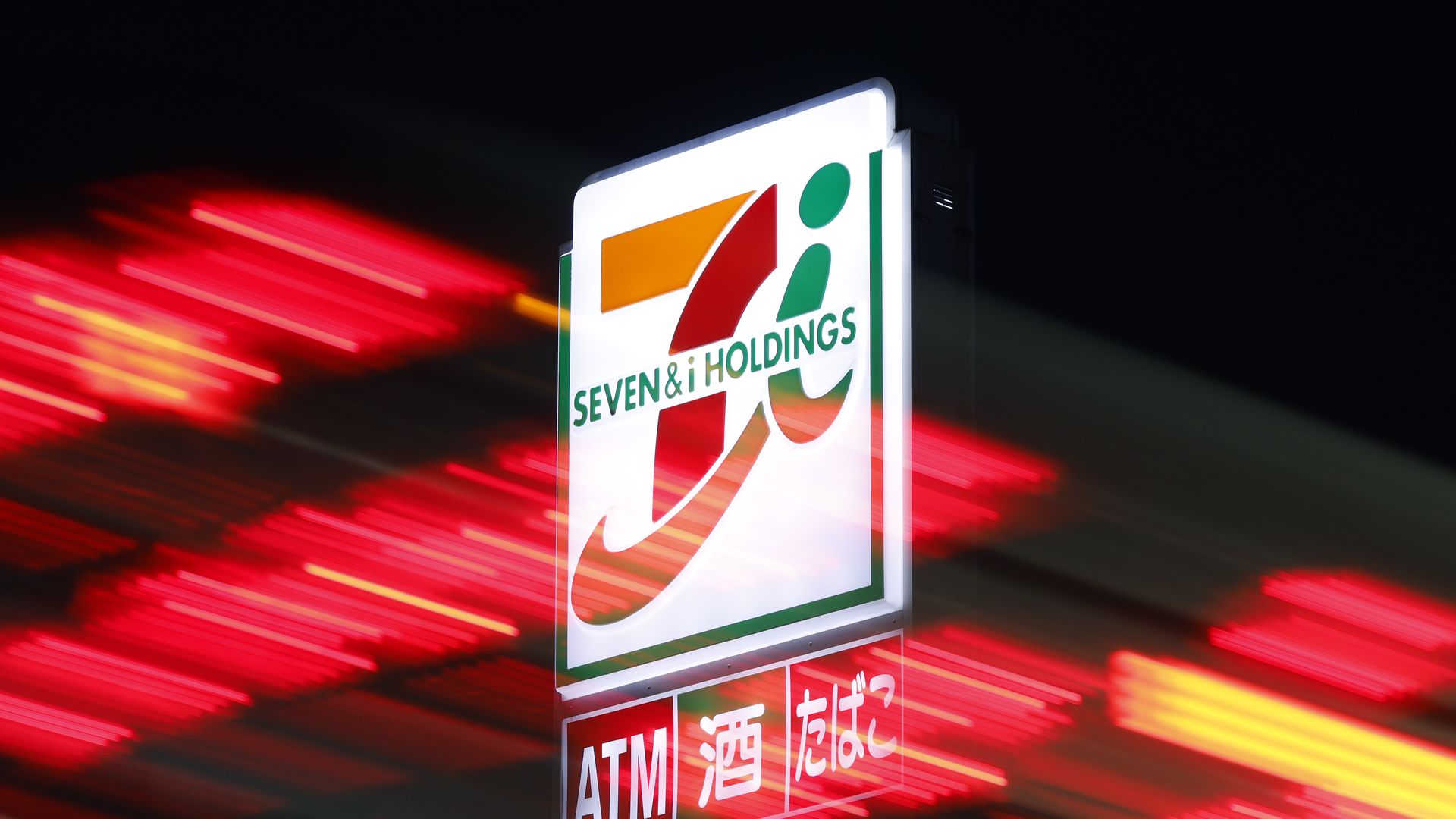 Illuminated Seven & i Holdings sign at night with blur red and yellow lights in the background, featuring ATM, 酒 (alcohol), and たばこ (tobacco) in Japanese.