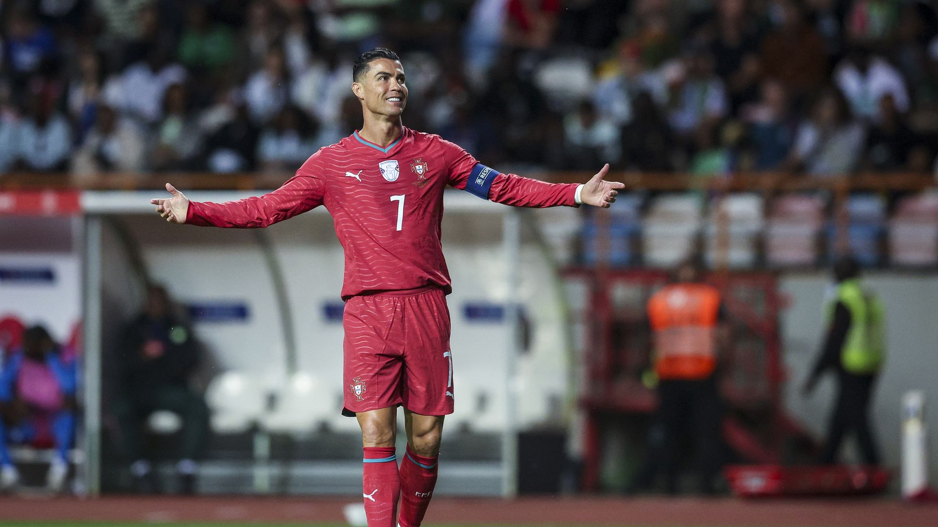 Cristiano Ronaldo in his red Portugal uniform holds his arms out and smiles while on the soccer pitch