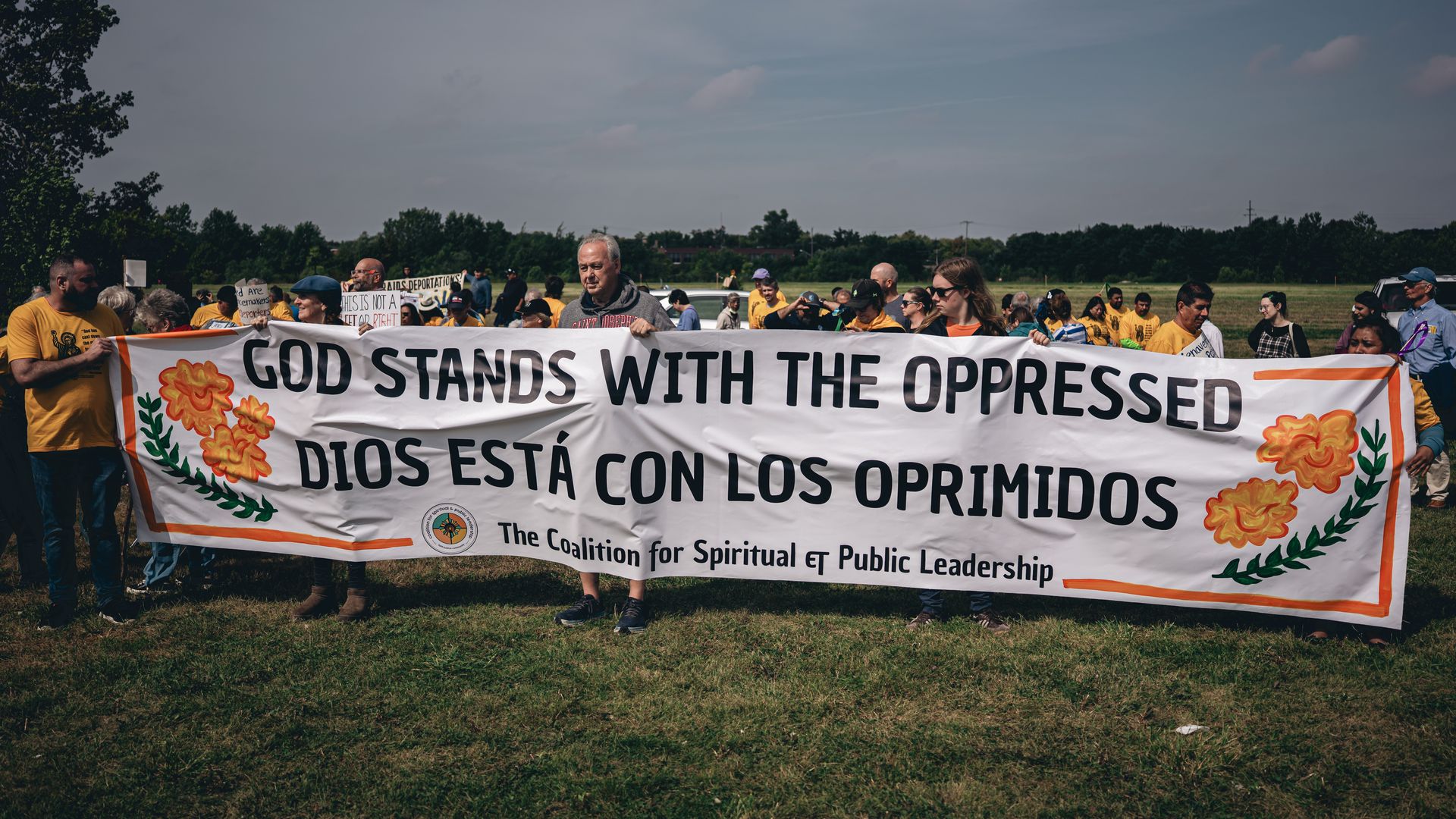 A diverse group of people outdoors holding a large white banner with orange flowers that reads, "GOD STANDS WITH THE OPPRESSED / DIOS ESTΓ CON LOS OPRIMIDOS," by The Coalition for Spiritual and Public Leadership.