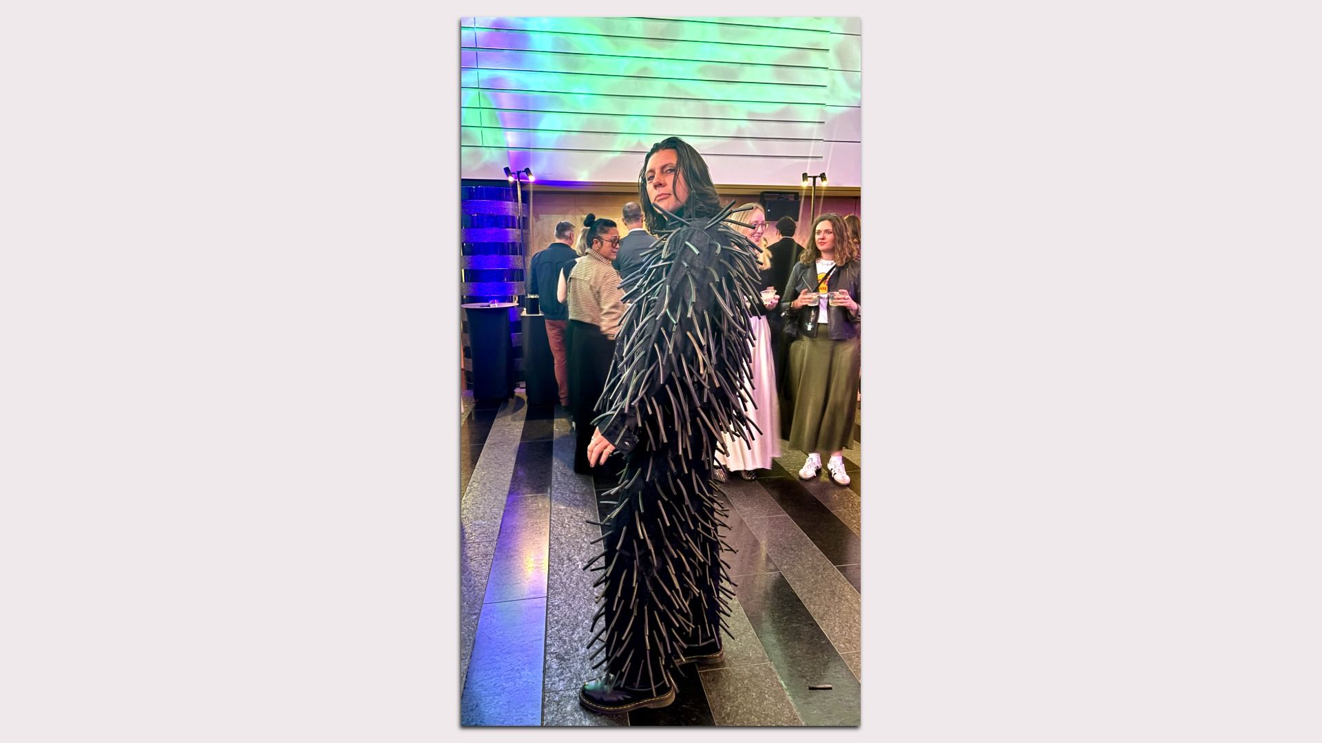 Person wearing a black spiky full-body outfit stands at a party; colorful neon lights and guests fill the background.