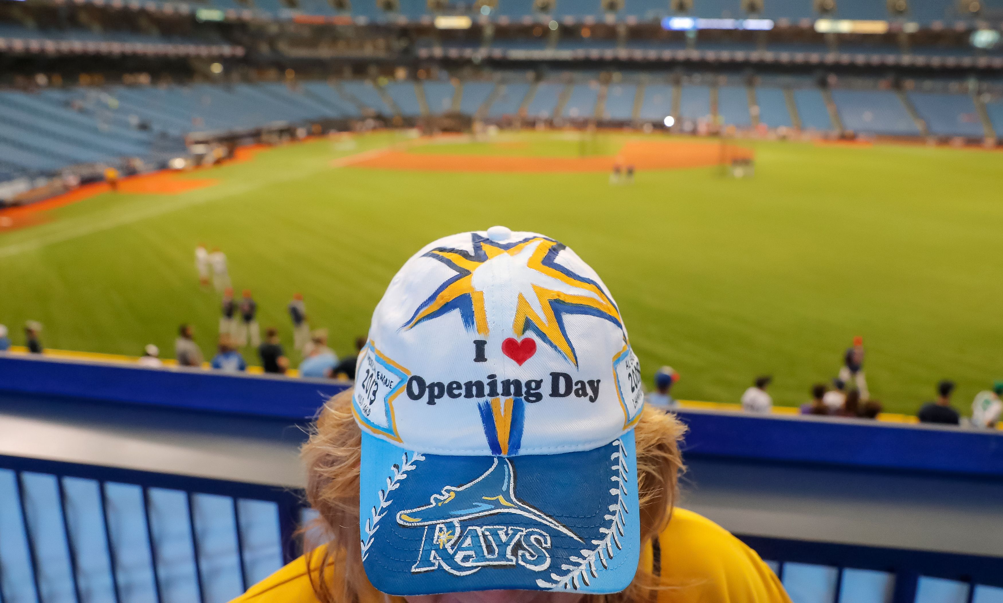 A person standing in front of the field at Rays stadium puts their head down to show their hat that says "I (heart) opening day" with the Rays logo on it