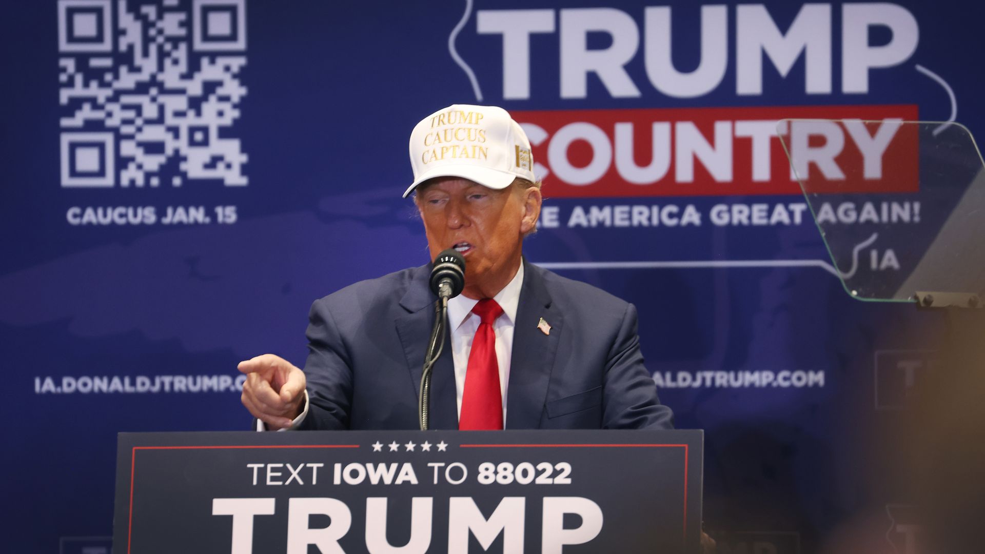 Donald Trump speaks in front of a sign that says "Trump Country" promoting the Iowa caucuses. He is pointing in front of him. He's wearing a suit with a red tie and a white cap. 