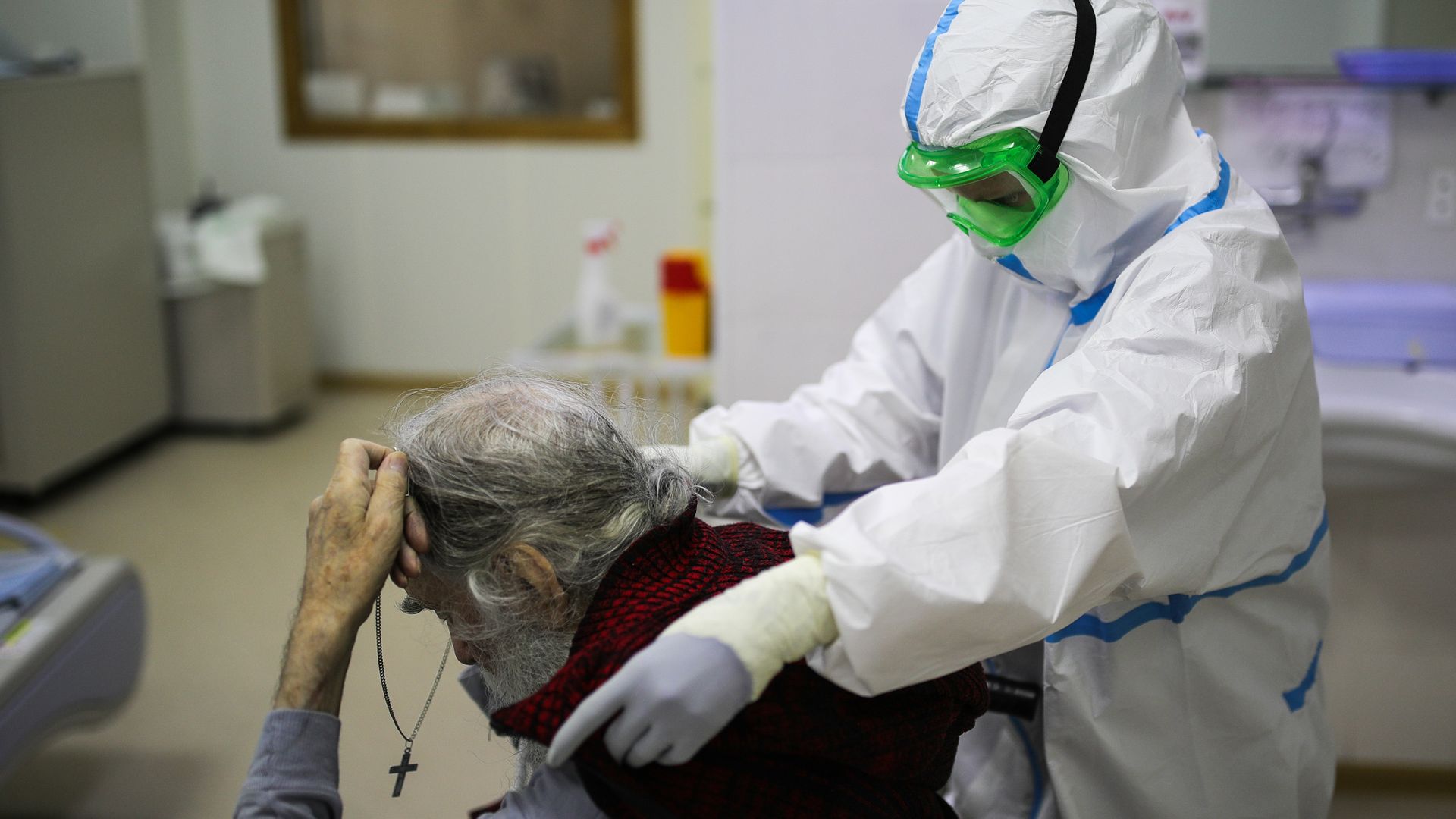 In this image, a person in a hazmat suit puts a blanket around the shoulders of a man holding a cross