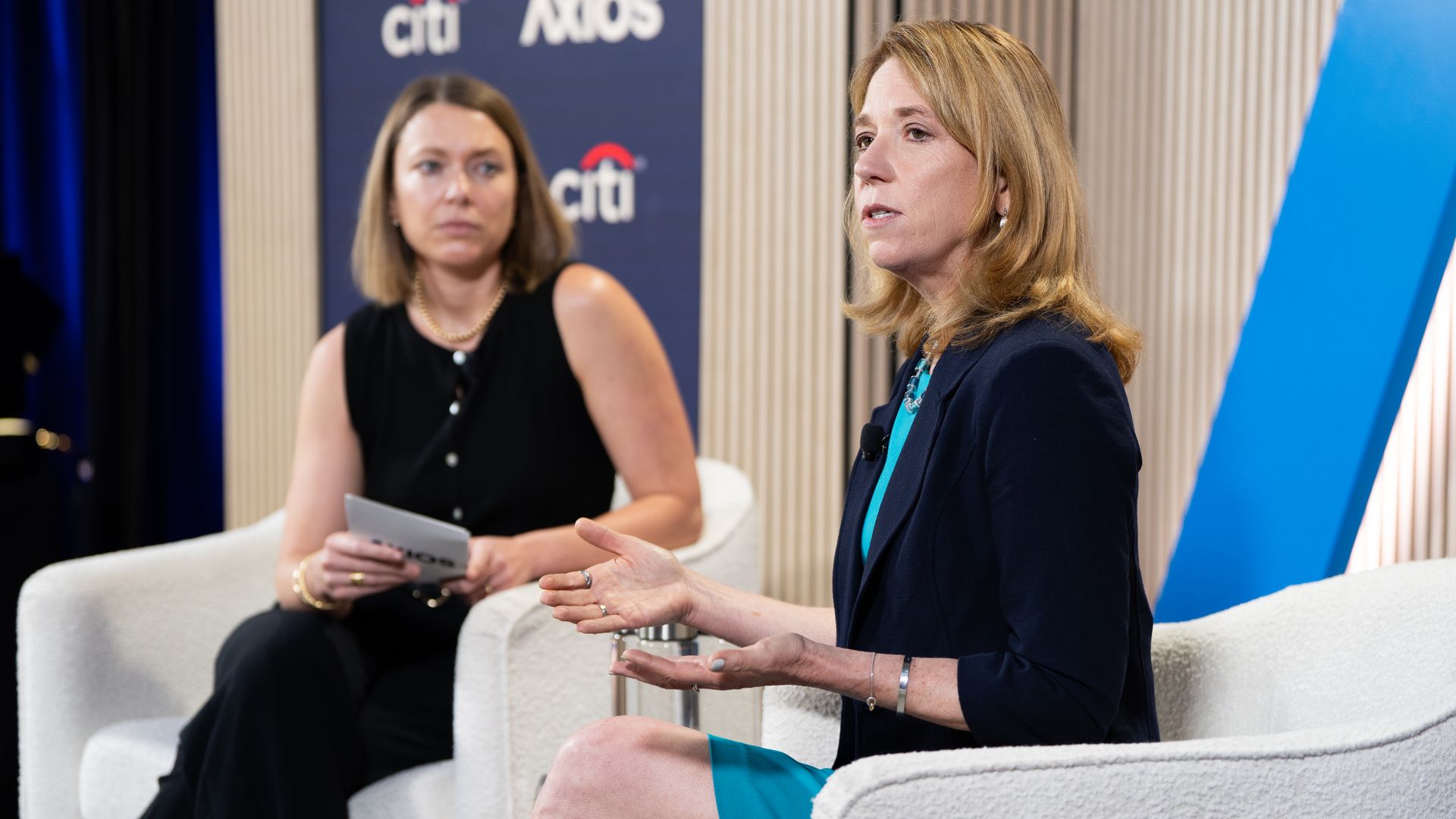 Two women in a panel discussion. The blonde on the right wears a navy blazer and turquoise dress and speaks, while the other in black holds notes; a blue Citi/Axios backdrop behind them.
