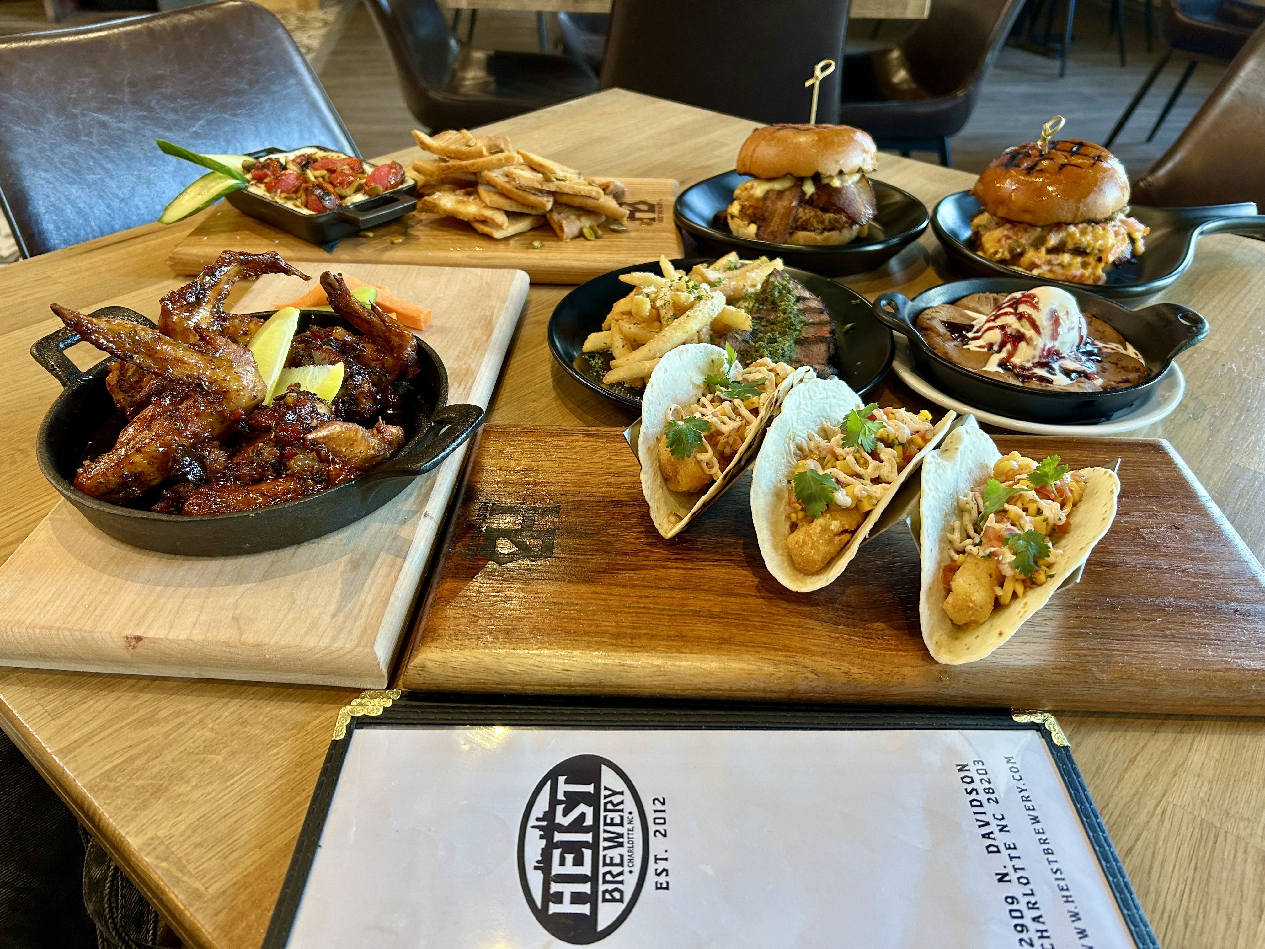 heist brewery. Table displays a spread: skillet of glazed ribs with lemon, board of fries, two burgers on black plates, a pasta dish, soft tacos, and a skillet dessert with vanilla ice cream and berry sauce.