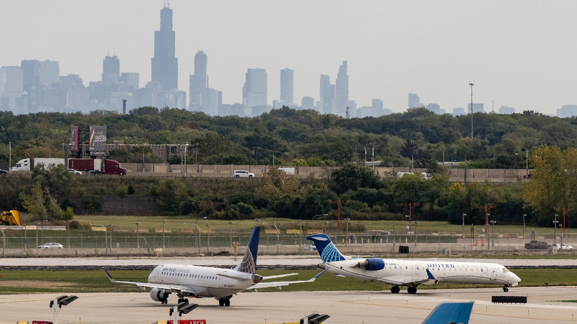 Two United Express airplanes taxiing on a runway with green trees and highway in the background, and a hazy city skyline with tall buildings visible beyond the trees.