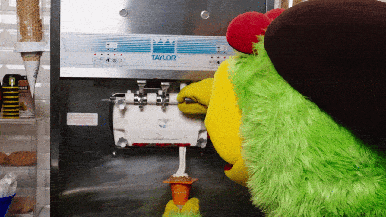 Close-up of a bright green fuzzy puppet with a yellow beak and red crest pressing a lever on a Taylor beverage machine; chrome controls and an orange cone component are visible.