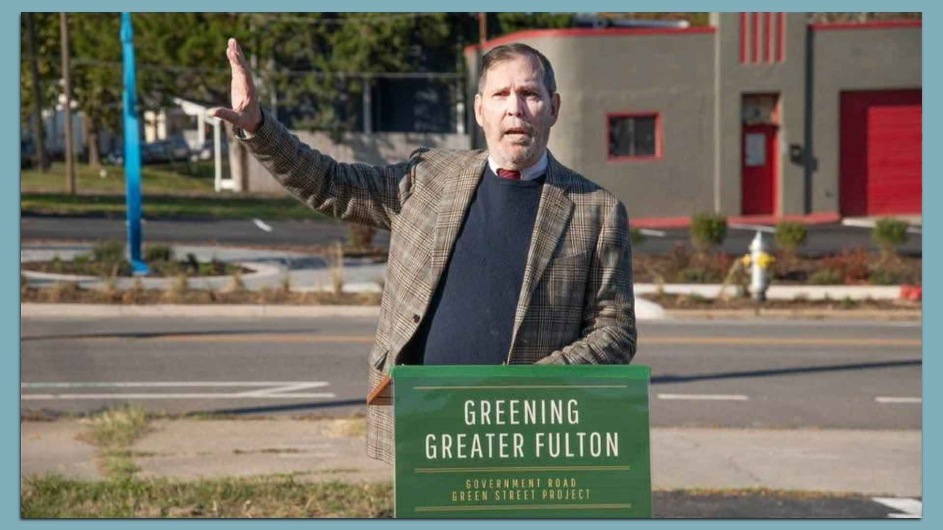 Middle-aged man in a brown plaid blazer and navy sweater raises his left arm on a street. A green sign in front reads "Greening Greater Fulton" with a gray building and red doors in the background.