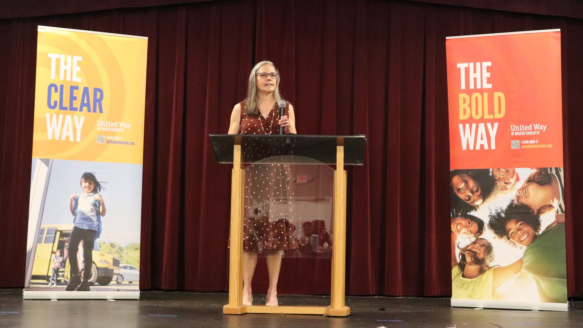 A speaker addresses an audience from a podium at a United Way event, flanked by banners reading "The Clear Way" and "The Bold Way."
