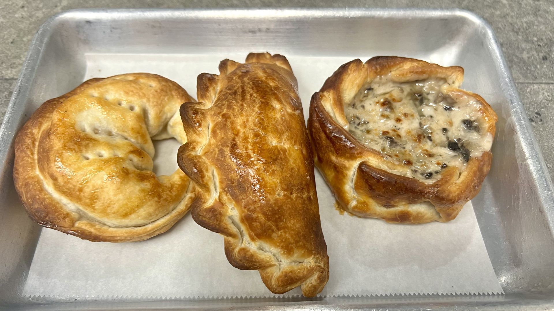 Three golden-brown baked pastries on parchment paper in a metal tray: a crescent-shaped pastry, a crimped half-moon-shaped pastry, and a round pastry with a creamy mushroom filling.