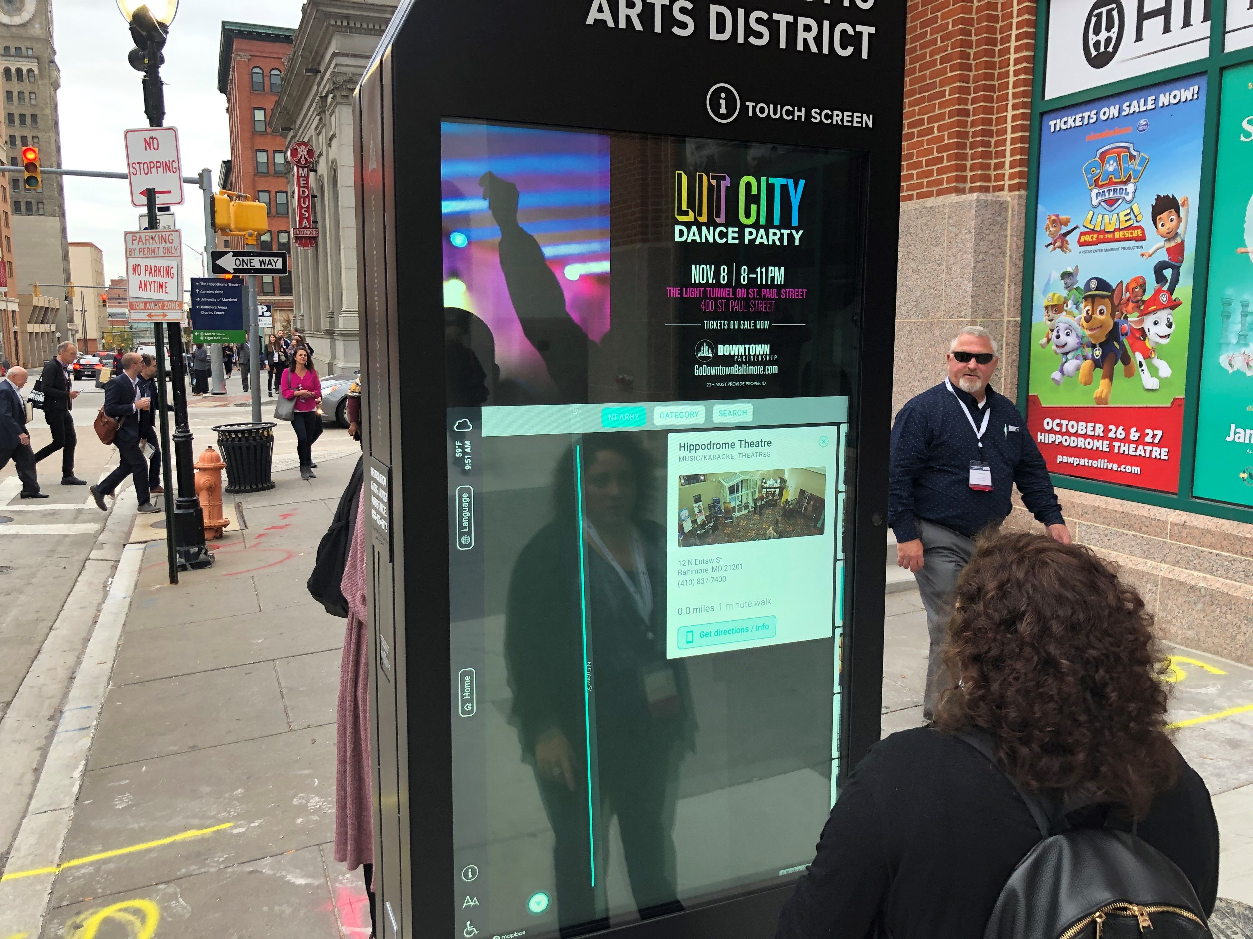 A person uses one of the touch-screen kiosks in another city, looking at digital content while people walk by on the sidewalk.