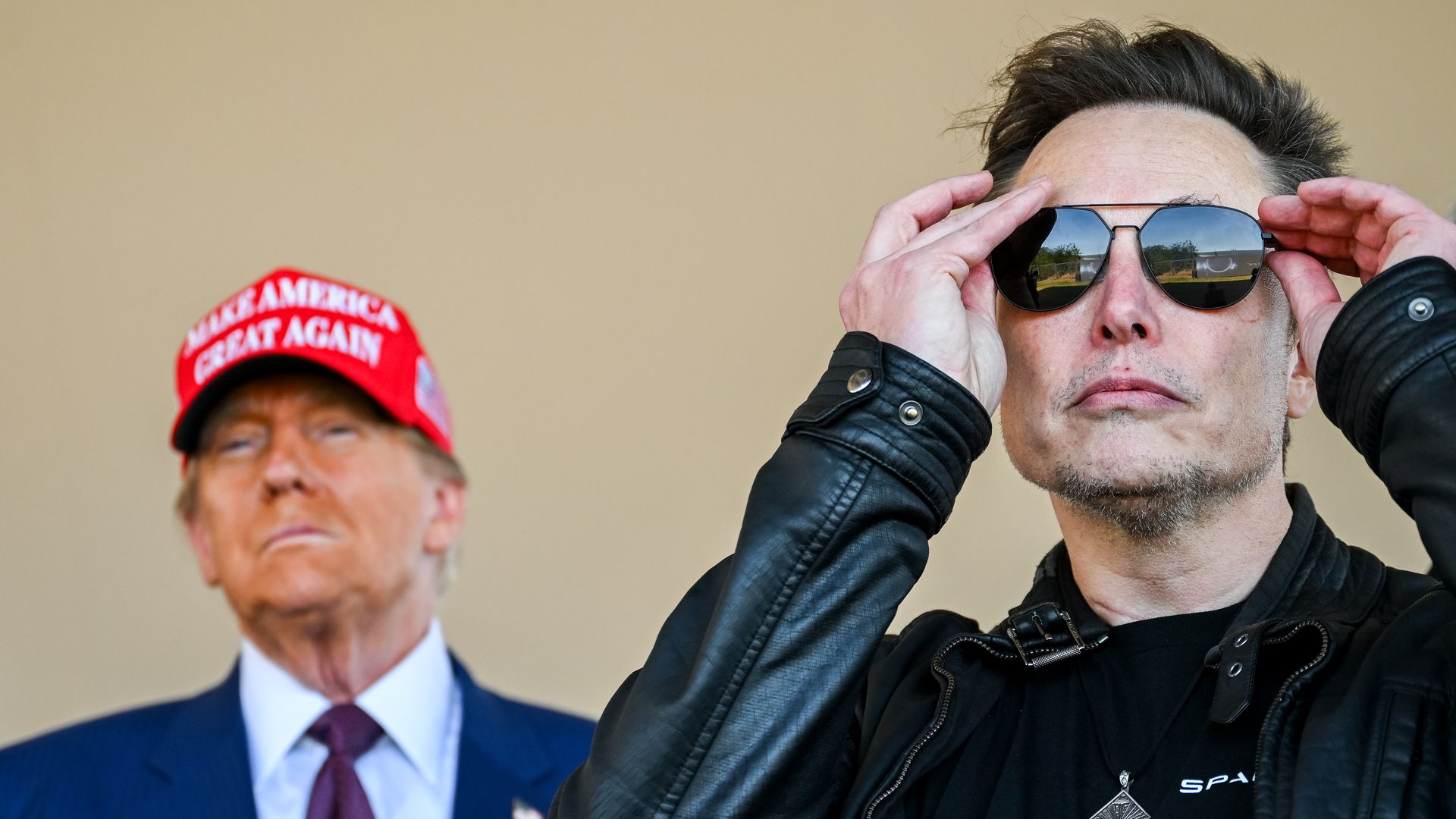 Trump wearing a MAGA hat behind Elon Musk, putting on sunglasses
