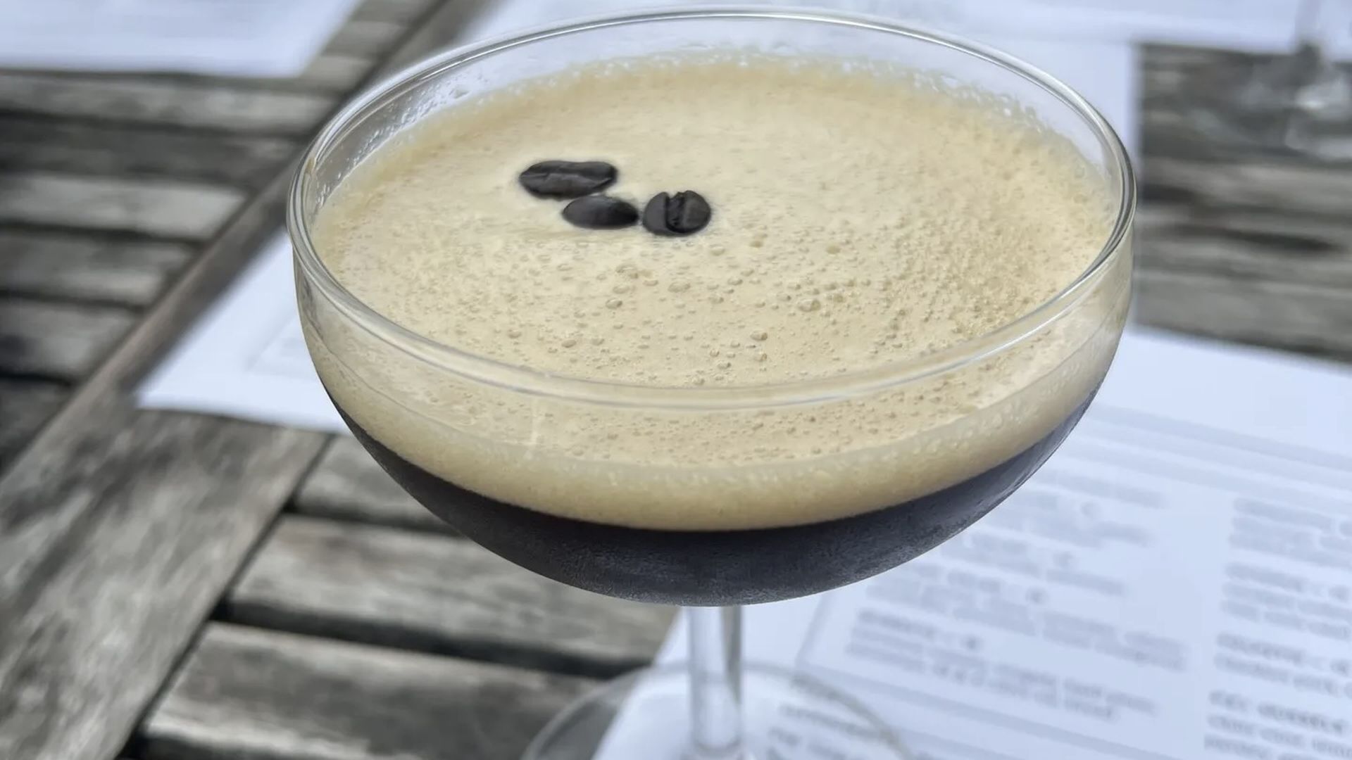 A coupe glass holds a frothy tan espresso martini with a dark bottom layer, garnished with two coffee beans; sits on a weathered wooden table with papers nearby.