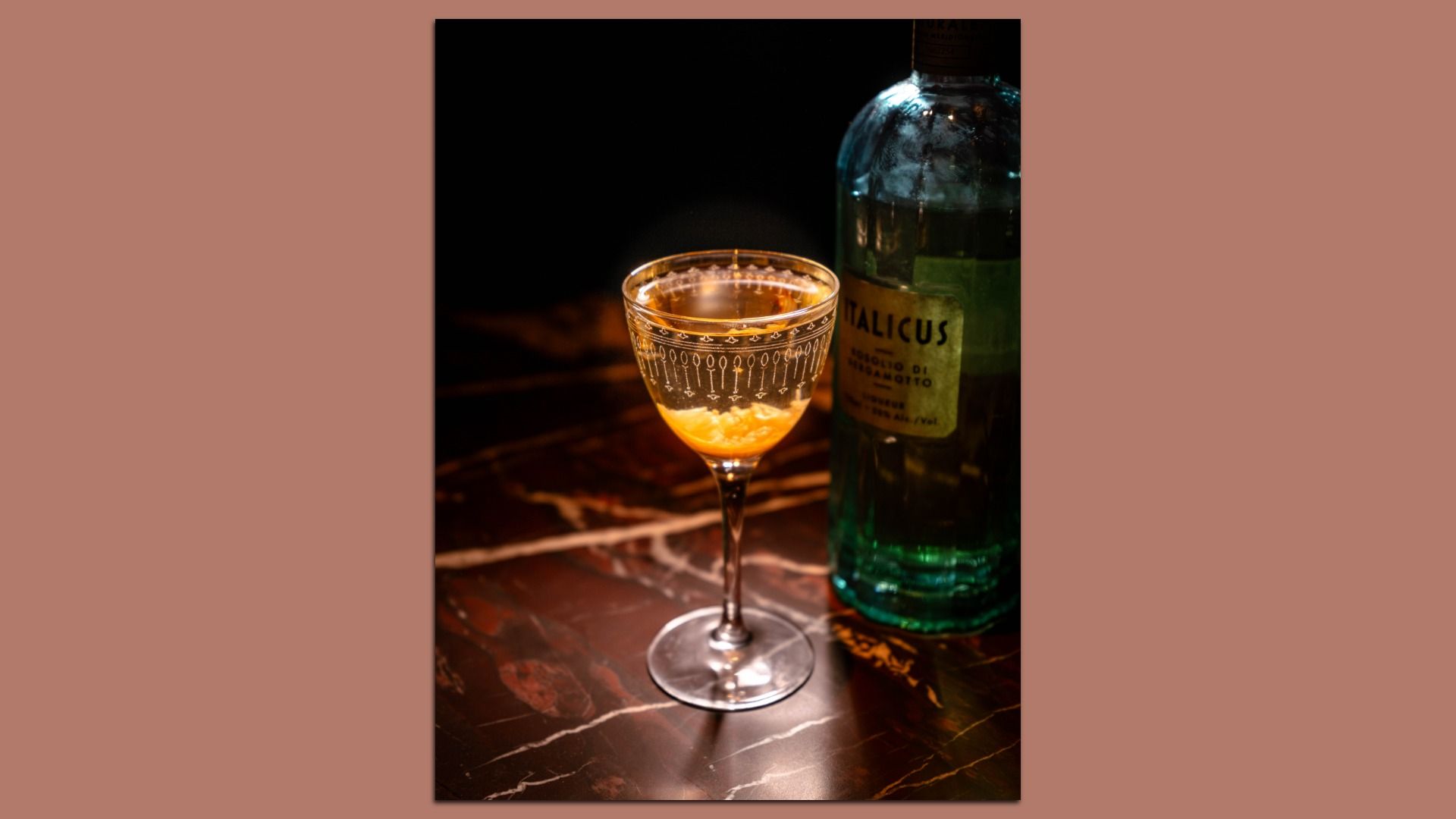 A glass with a golden cocktail on a dark marble surface next to a green glass bottle labeled Italicus, illuminated against a black background.