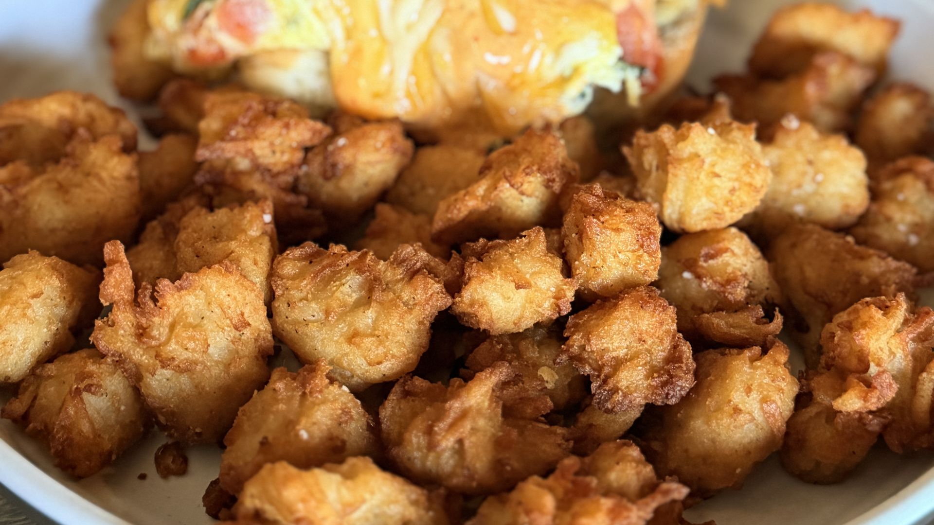 Plate of golden, crispy fried chicken bites; in the background a large fried sandwich with melted cheese and greens, resting on the plate.