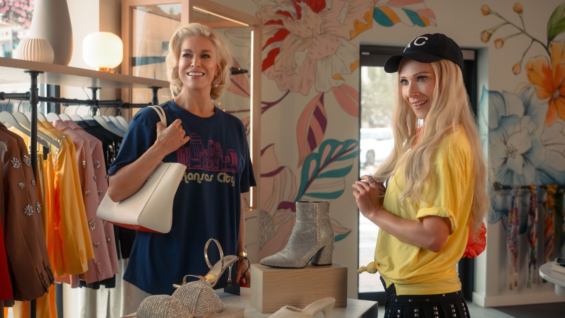Two smiling women in a bright boutique browsing clothes; one wears a blue Kansas City T-shirt and carries a white bag, the other a yellow top with a black cap. Colorful racks and a floral mural.