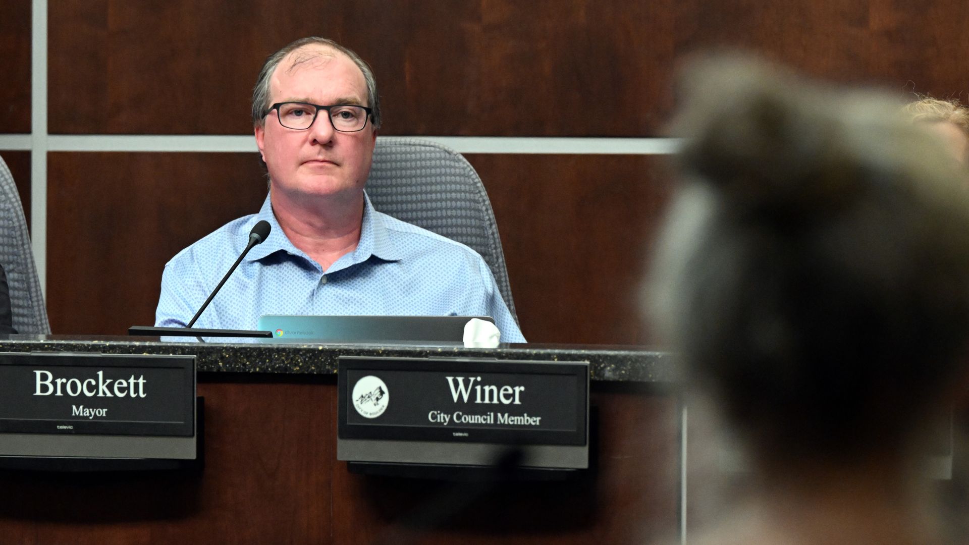 Boulder Mayor Aaron Brockett seated behind a desk with nameplate "Brockett, Mayor" in a wood-paneled room, looking forward at an out-of-focus person in front.