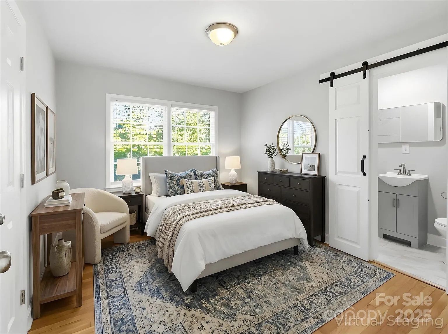 Bright bedroom with white walls, queen bed with white and blue bedding, wooden dresser with round mirror, beige chair, wooden side table, sliding barn door leading to bathroom.