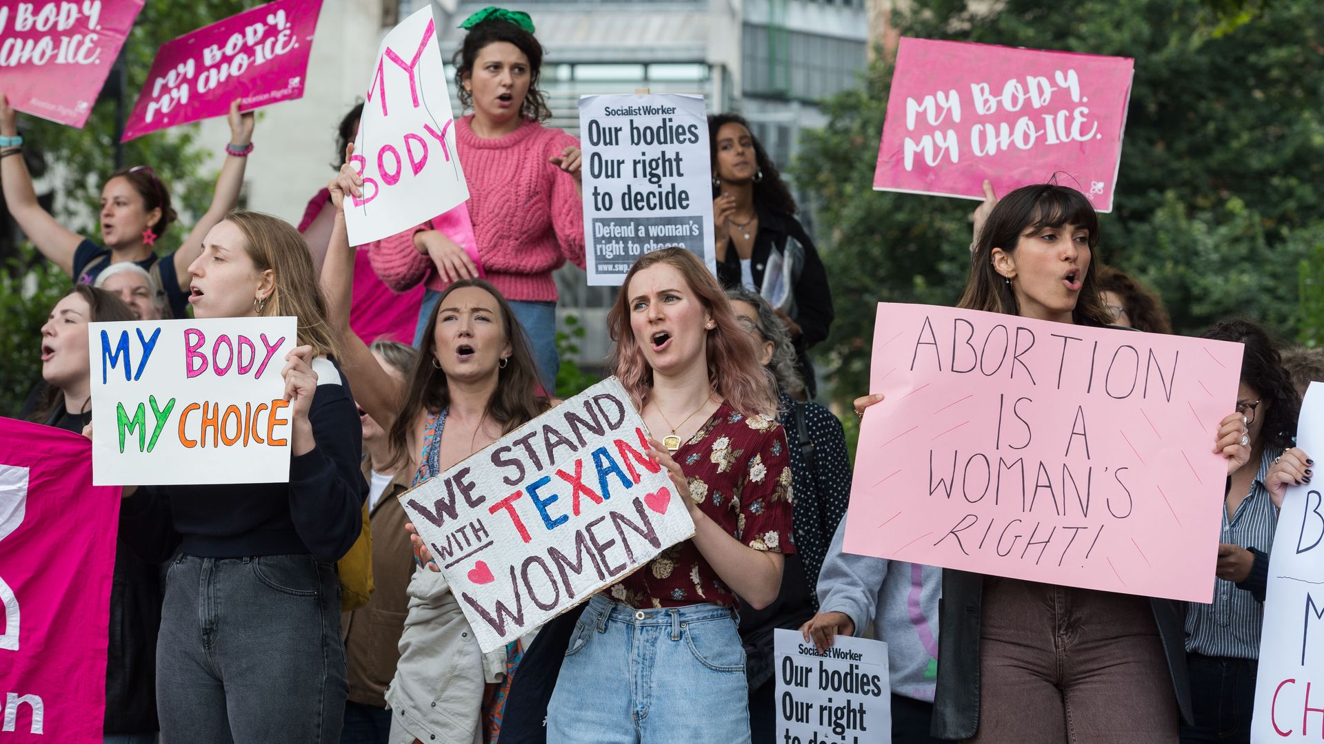 A group of women hold pink and white signs protesting texas abortion ban at an outdoor gathering.
