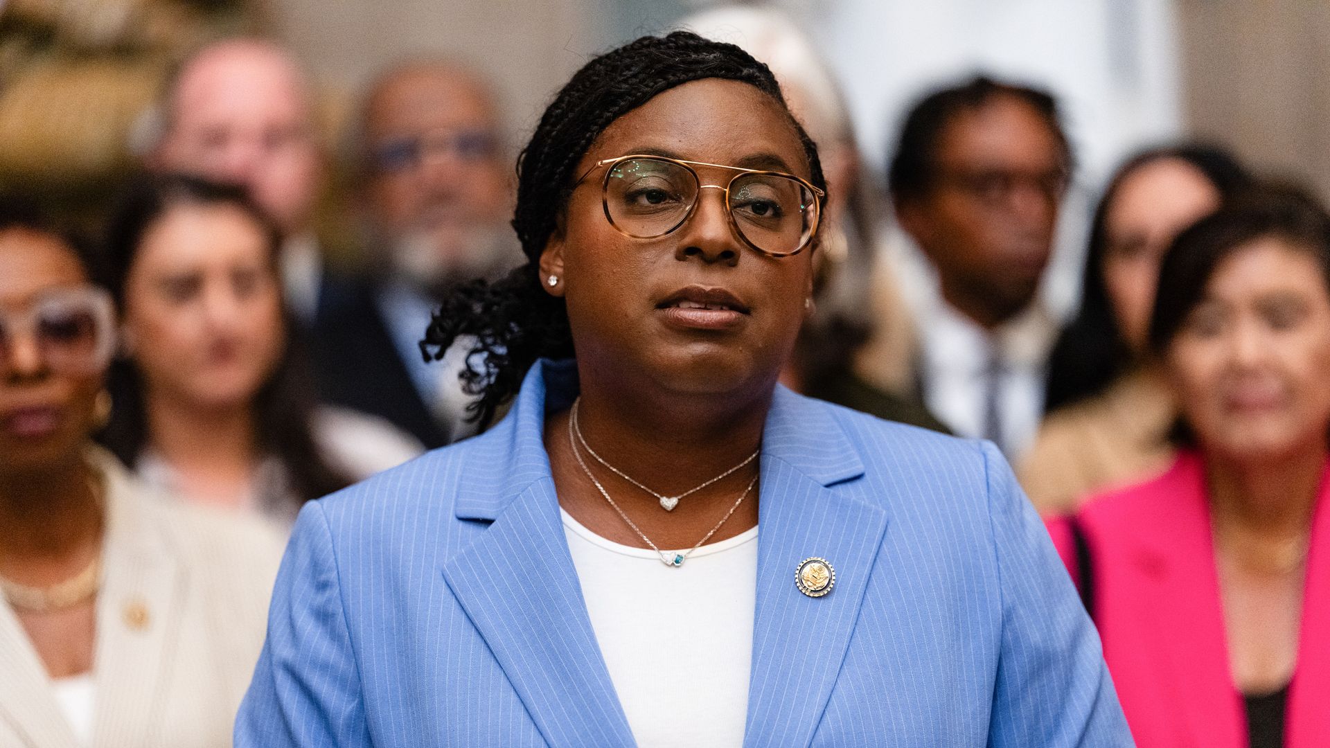 Person wearing a light blue pinstripe blazer and glasses speaking, with a blurred crowd behind them dressed in formal attire.