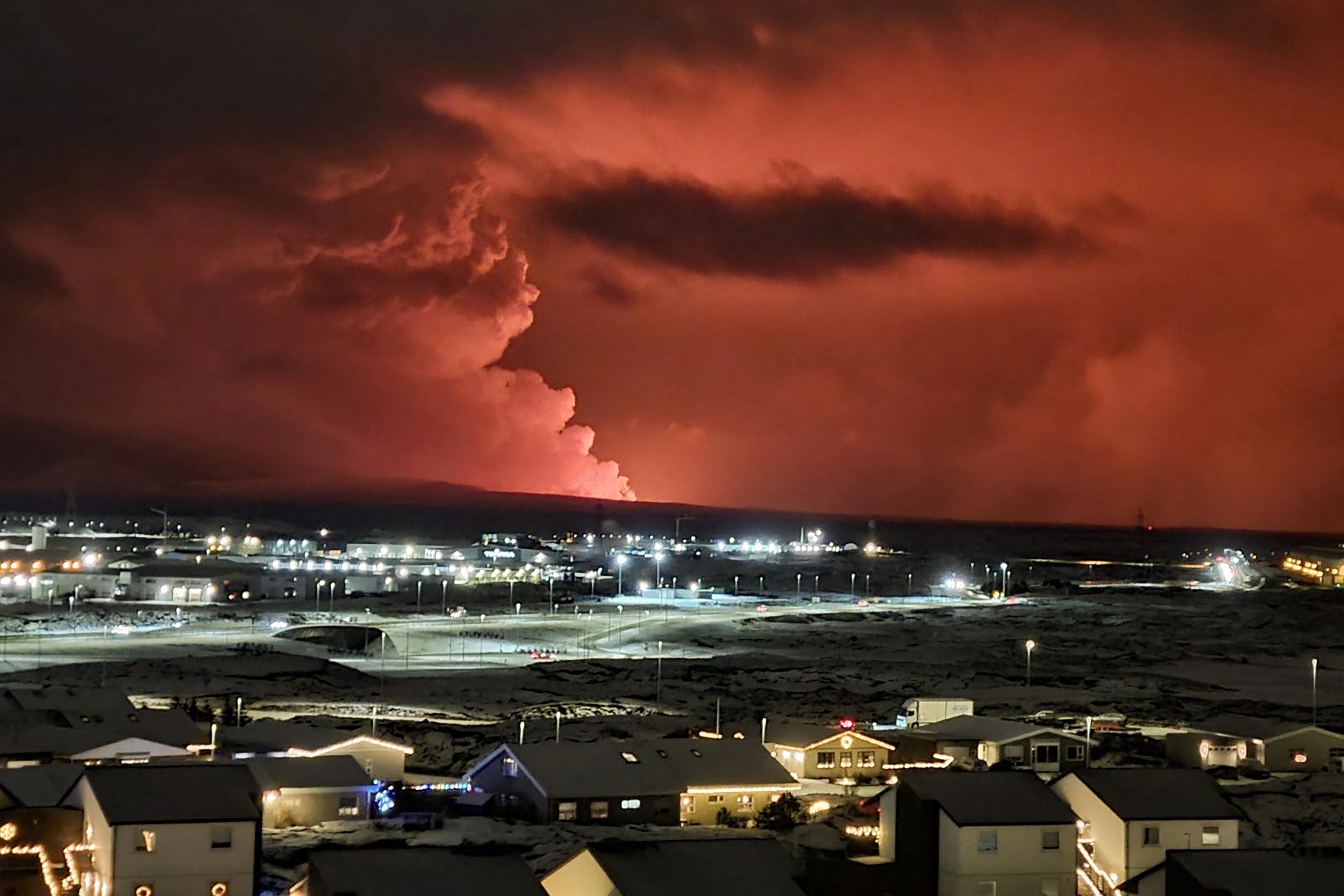 Houses in the village of Hafnarfjordur is seen as smoke is billowing in the distance as the lava colour the night sky orange from an volcanic eruption on the Reykjanes peninsula, western Iceland on December 18, 2023.
