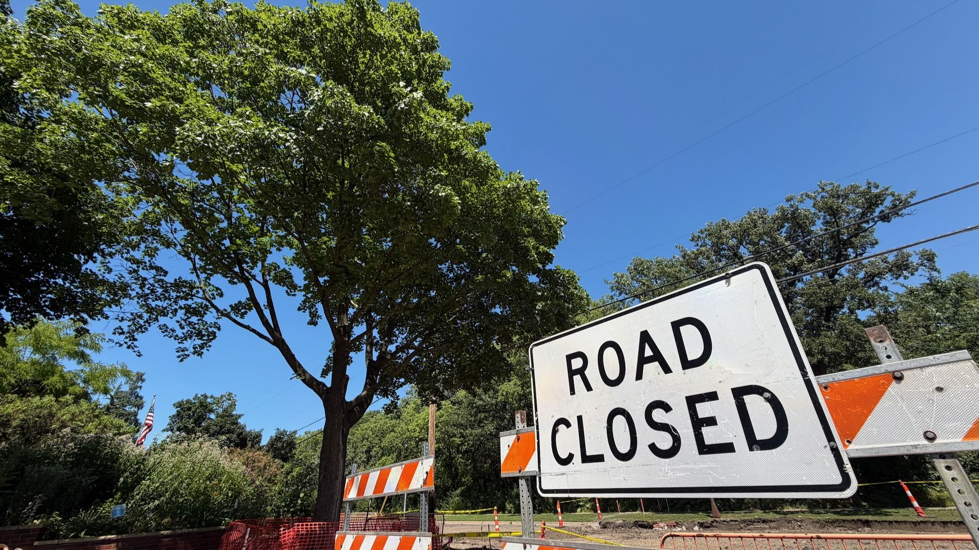Bright sunny day with a blue sky and green trees; orange construction barriers block a street, and a large "ROAD CLOSED" sign sits in the foreground.