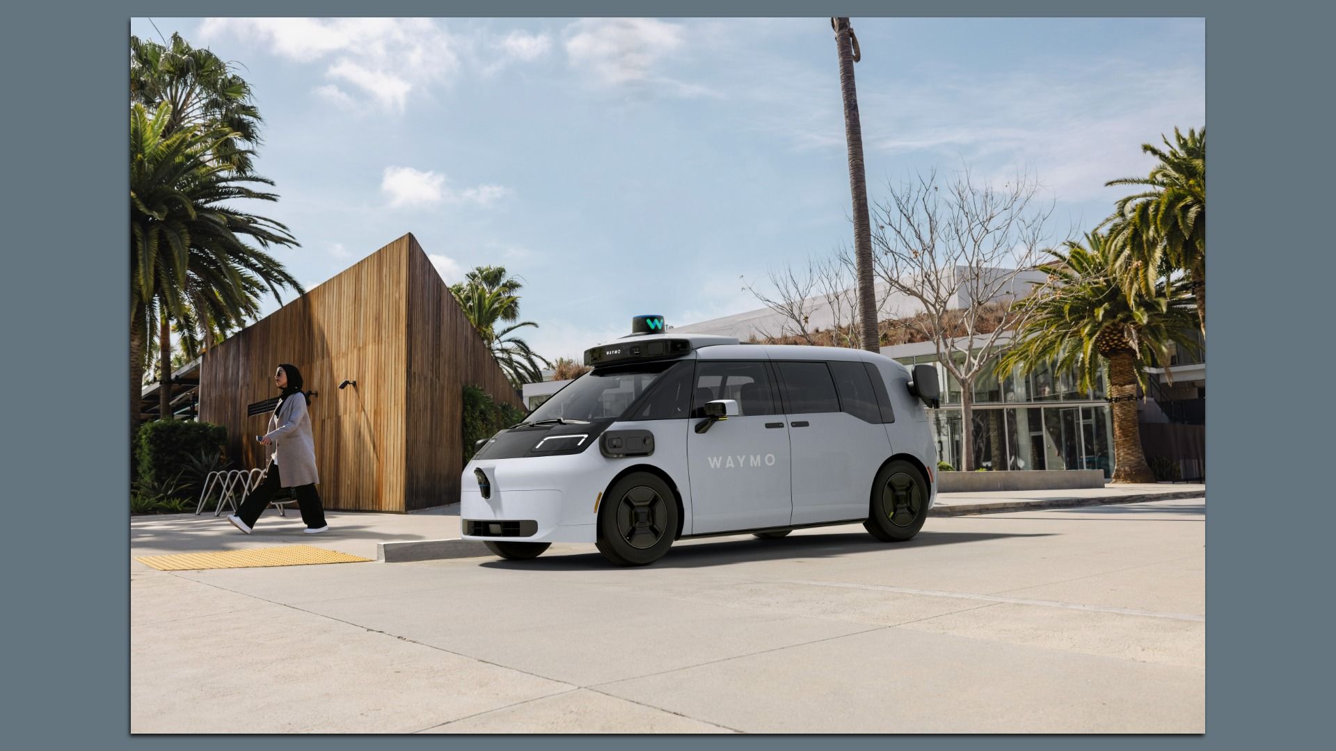 White Waymo autonomous minivan parked near a wooden building and palm trees, with a woman in a gray coat and black pants walking on the sidewalk. Bright sunny day with blue sky.