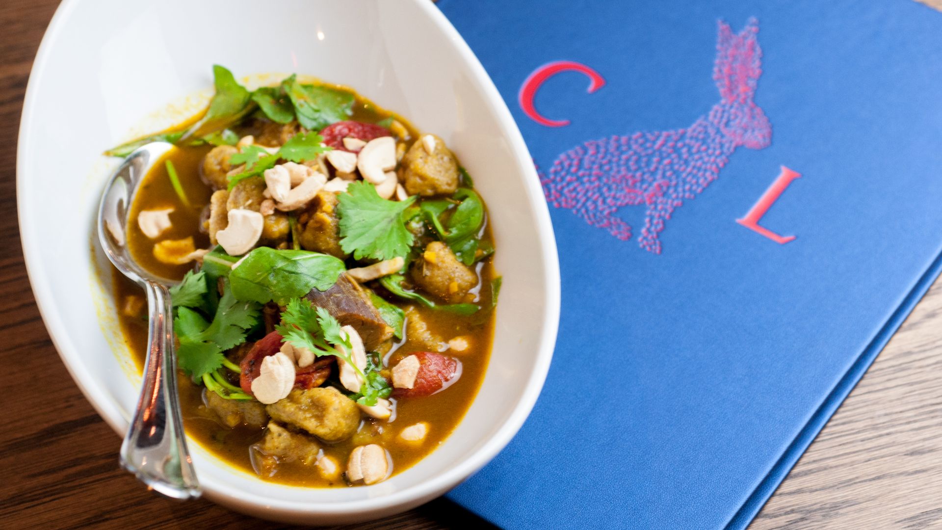 A white bowl of curried goat is topped with green garnish. A spoon pokes out of the bowl, and it sits on a menu book for Compere Lapin, which is embossed with a rabbit image.