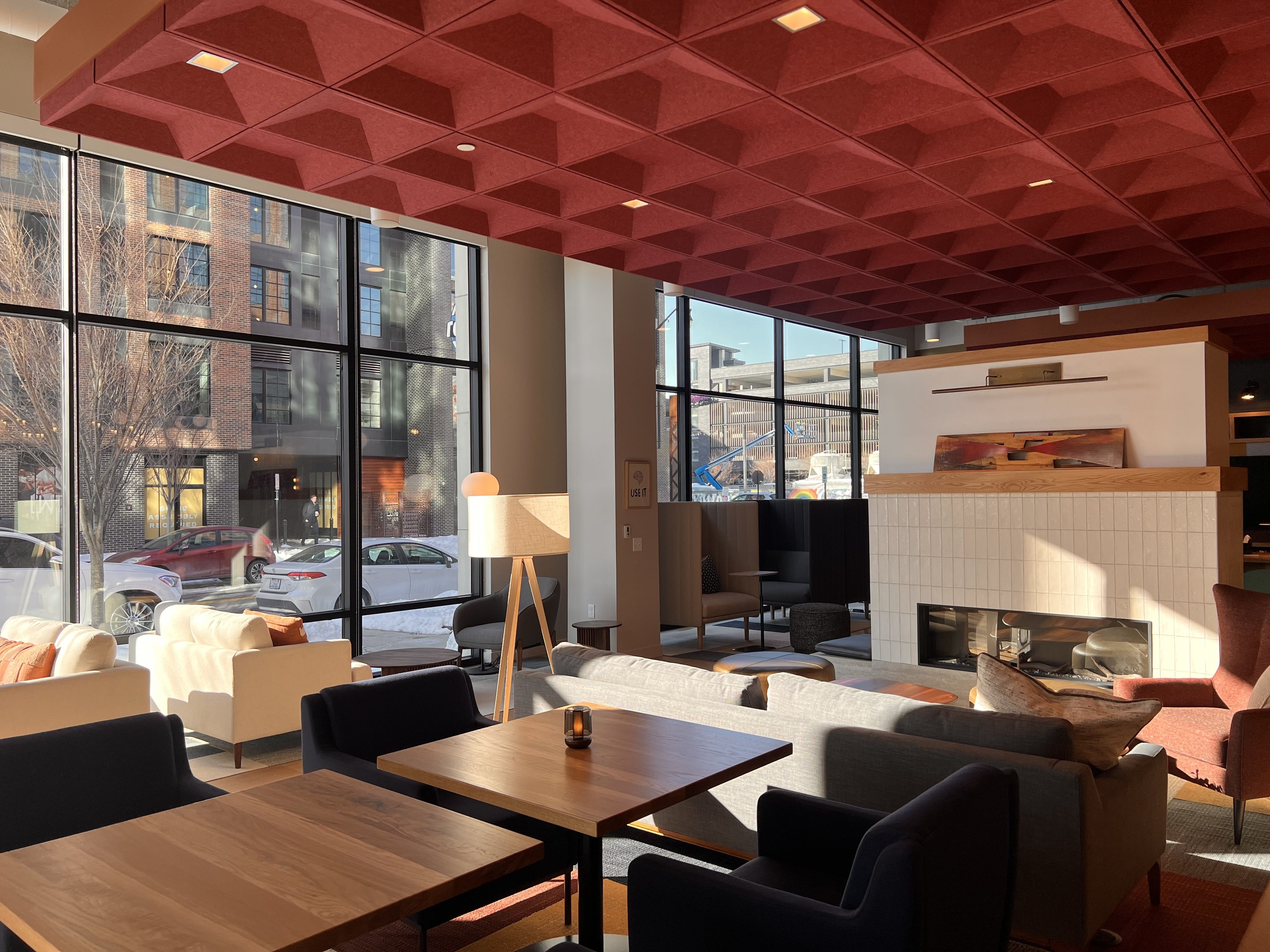 Modern lounge area with large windows, red textured ceiling, wooden tables, various chairs and sofas in neutral tones, and a white tiled fireplace with a colorful artwork above it.