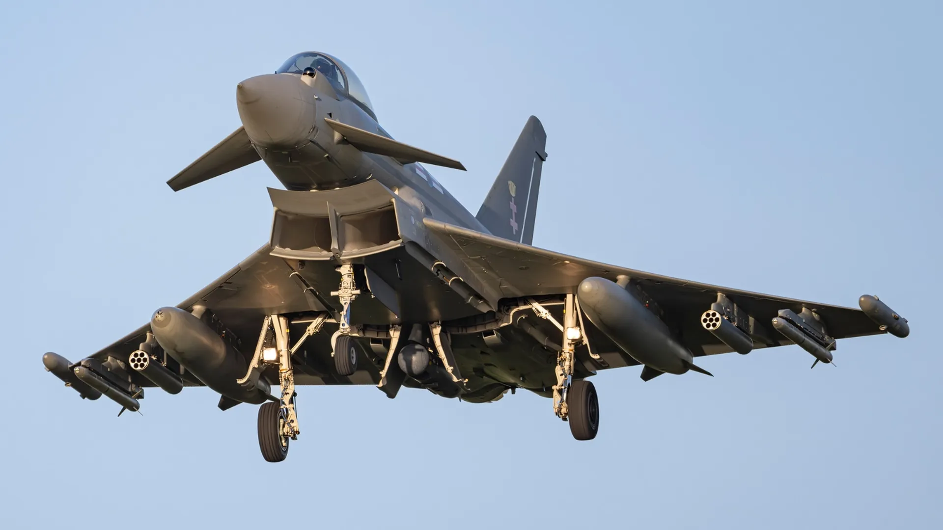 A gray military fighter jet in level flight with landing gear down, underwing missiles, bubble canopy, and a single tail, set against a clear blue sky.