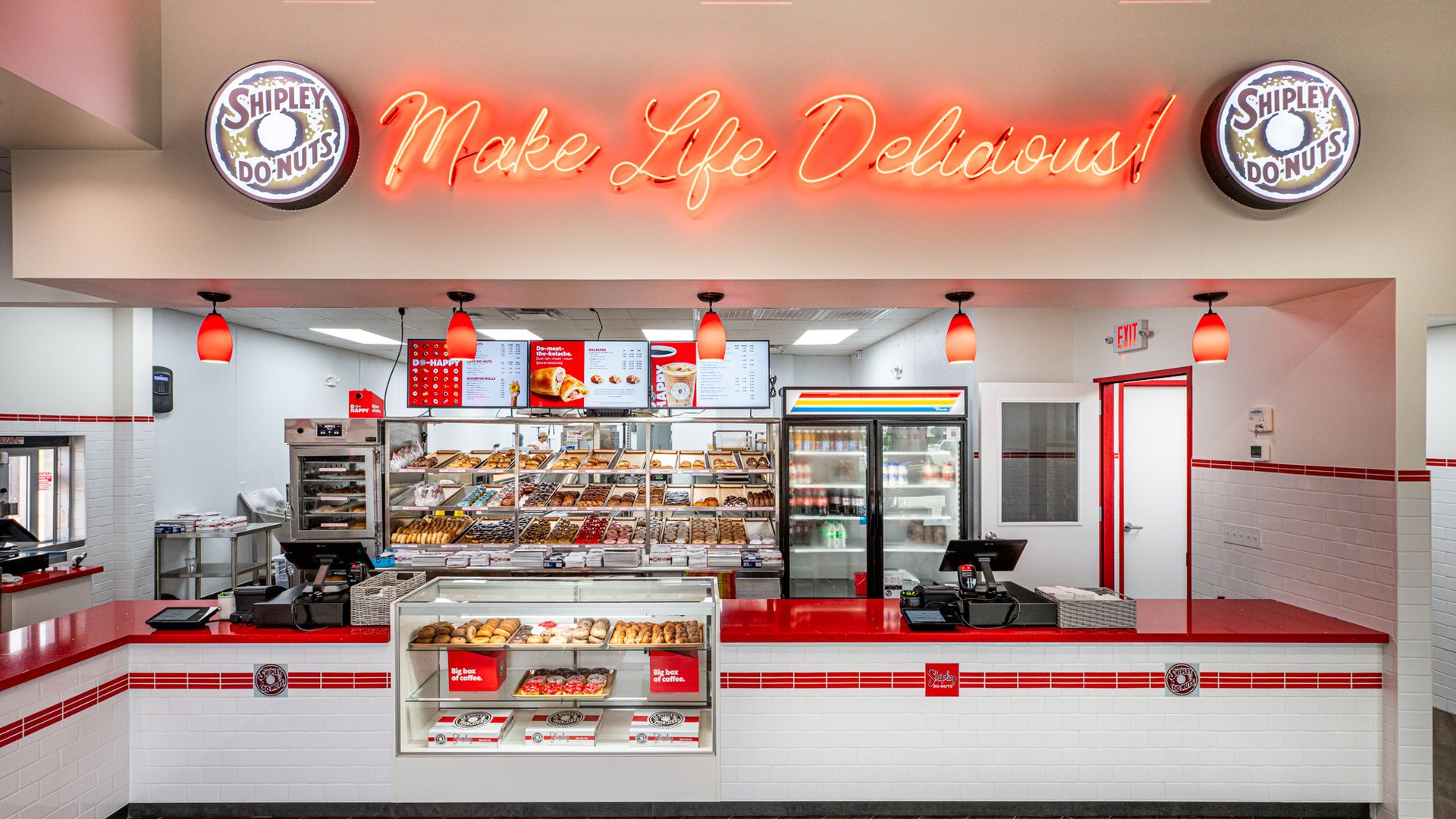Interior of a Shipley Do-Nuts shop: red neon sign "Make Life Delicious!" between circular logos; glass pastry display case and red-and-white counter.