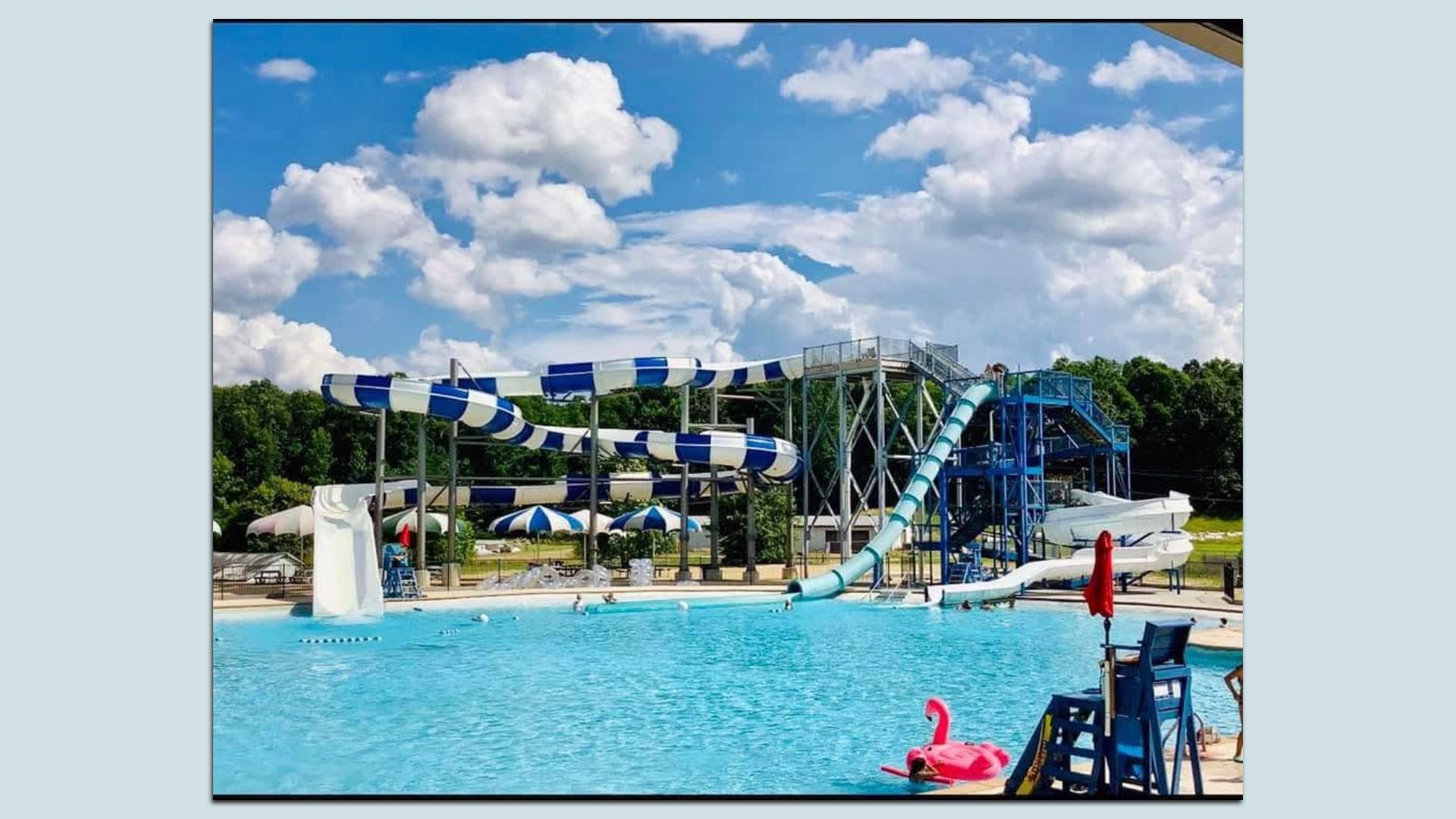 Outdoor water park with blue-and-white striped twisting slides, metal supports, and a large pool. Bright blue sky with fluffy white clouds and green trees in the background.