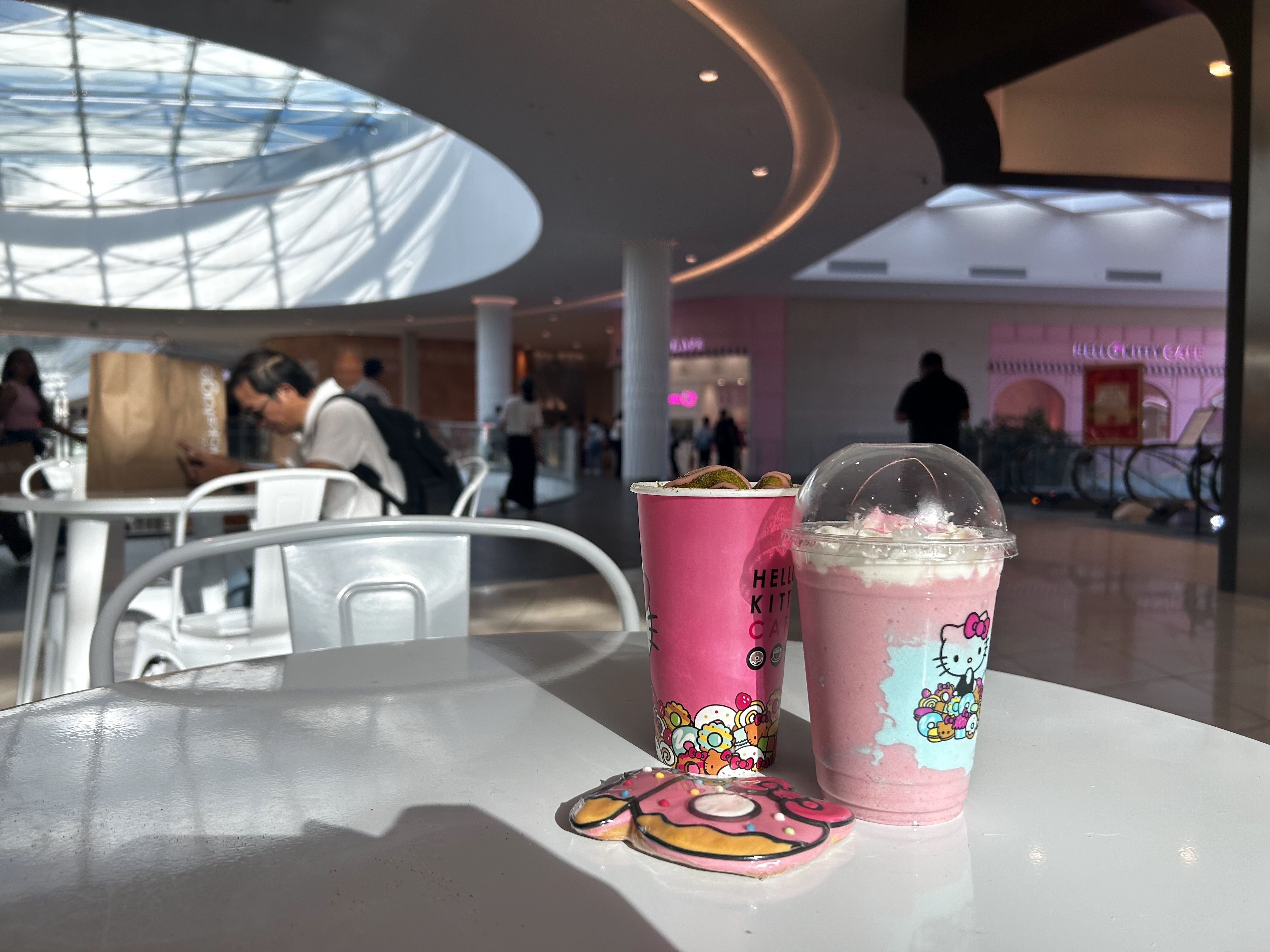 Two pink Hello Kitty drinks and a decorated pink cookie on a white table inside a mall cafe with people and a glass ceiling in the background.